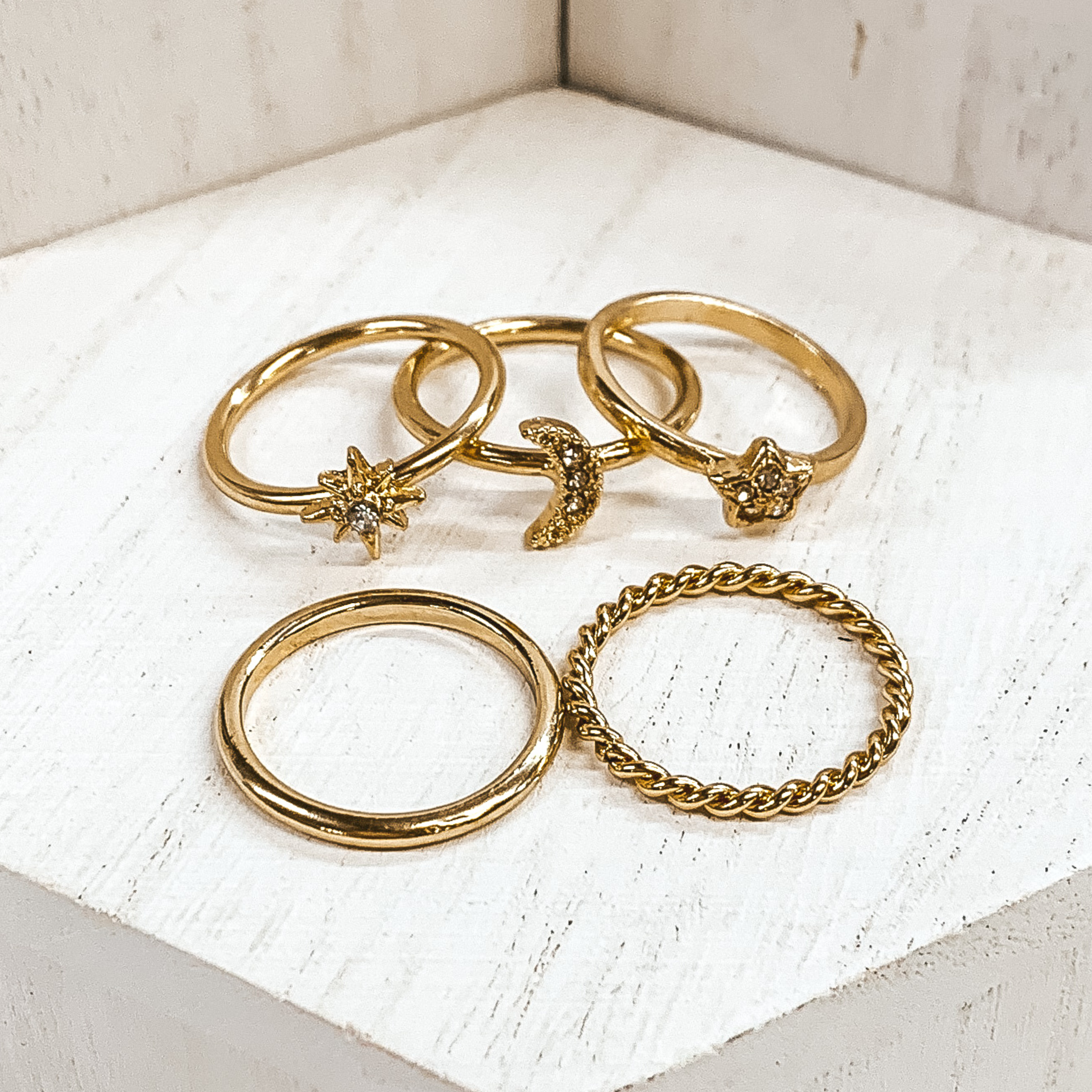 Set of five gold rings. One is a plain band and another is a twisted band.  Two rings have different star pendants, and the last ring has a moon pendant. These rings are pictured on white blocks. 