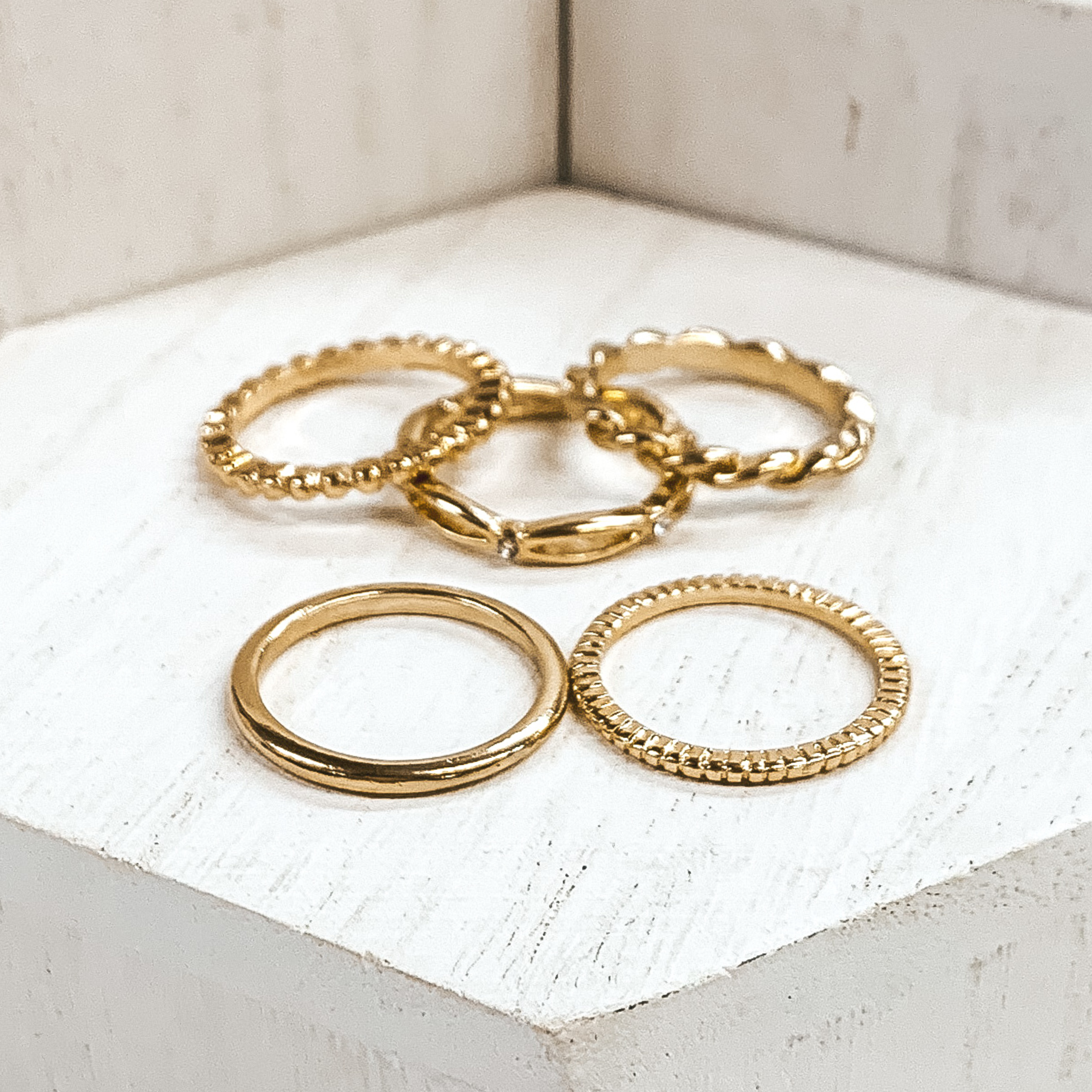 Set of five gold rings. One is a plain band, one is a bubble band, one is a twisted band, and the other two are textured bands. These rings are pictured on white blocks. 