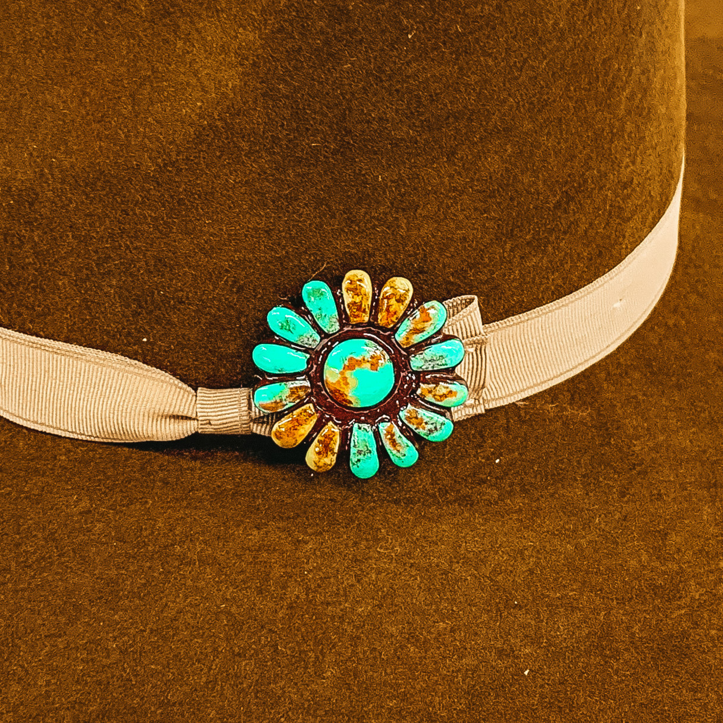 This hat pin is a flower concho. The main color is turquoise and is mixed with brown, red and yellow like real turquoise. This hat pin is pictured in front of a beige hat band.