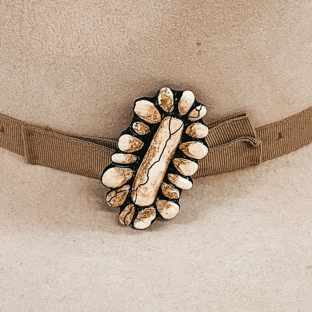This hat pin is a long, oval concho. The main color is ivory and is mixed with brown like real white buffalo. This hat pin is pictured in front of a beige hat band on a light colored hat.