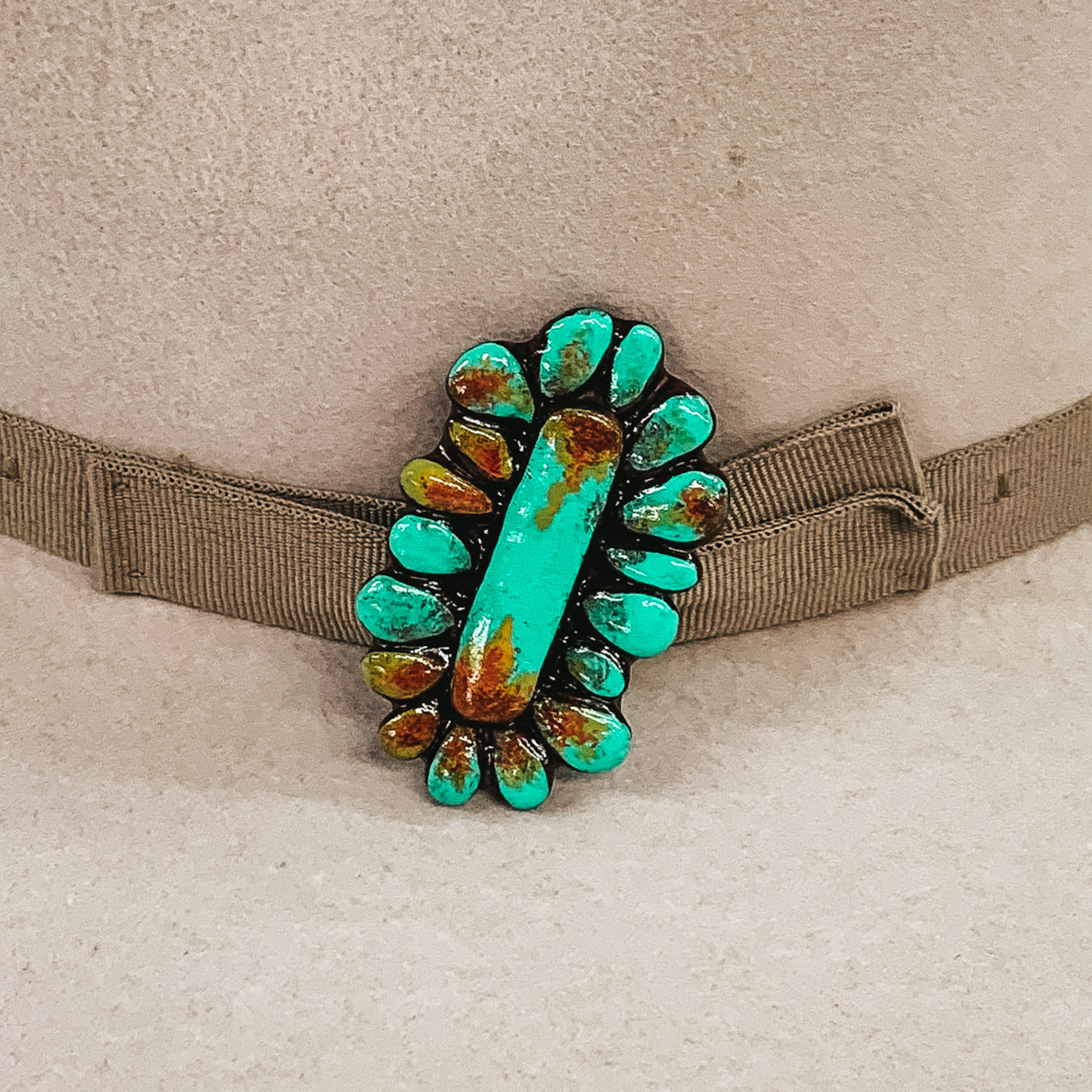 This hat pin is a long, oval concho. The main color is turquoise and is mixed with brown, red and yellow like real turquoise. This hat pin is pictured in front of a beige hat band on a light colored hat.