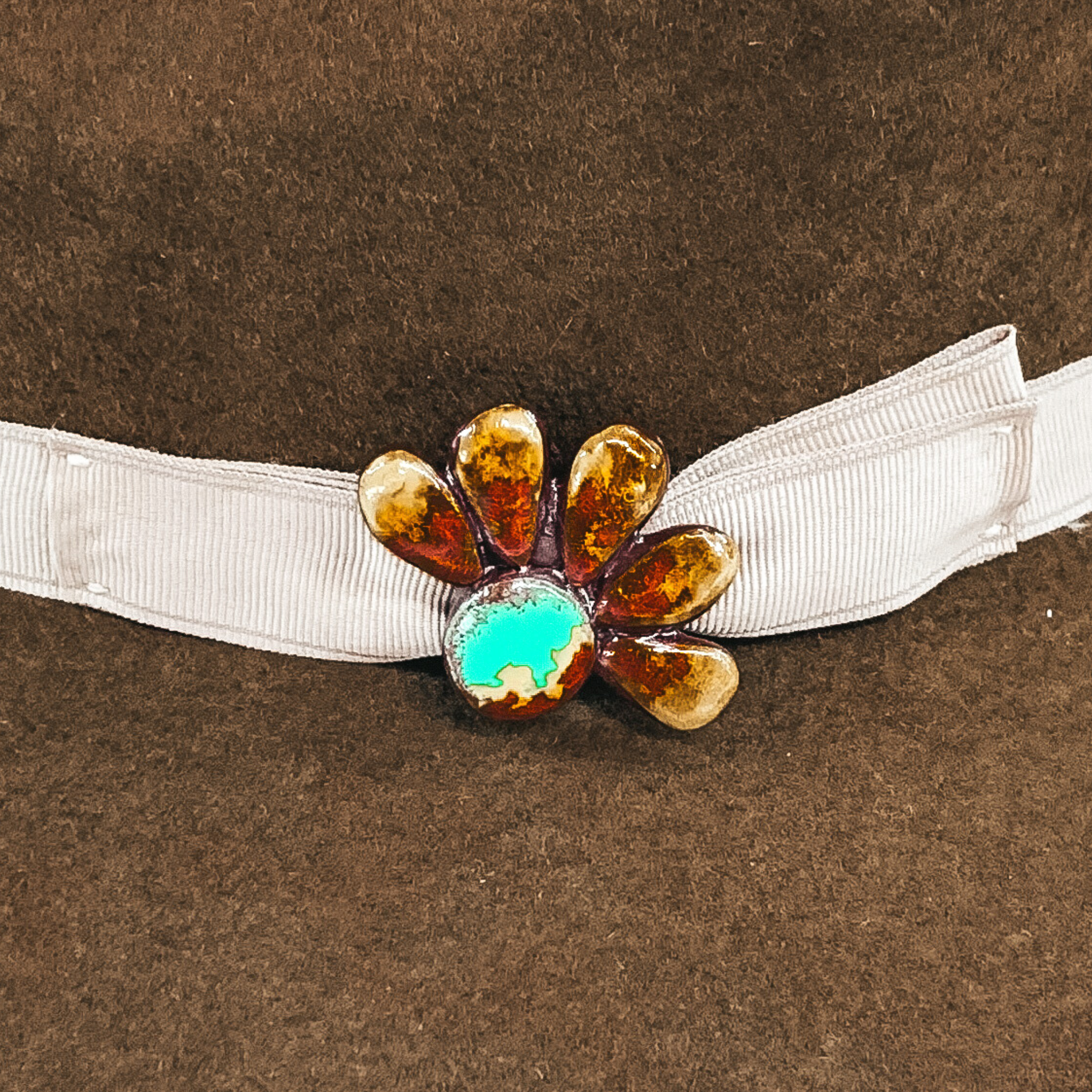 This hat pin is a half of a concho flower. The main color is turquoise and is mixed with brown, red and yellow like real turquoise. This hat pin is pictured in front of a beige hat band on a brown hat.