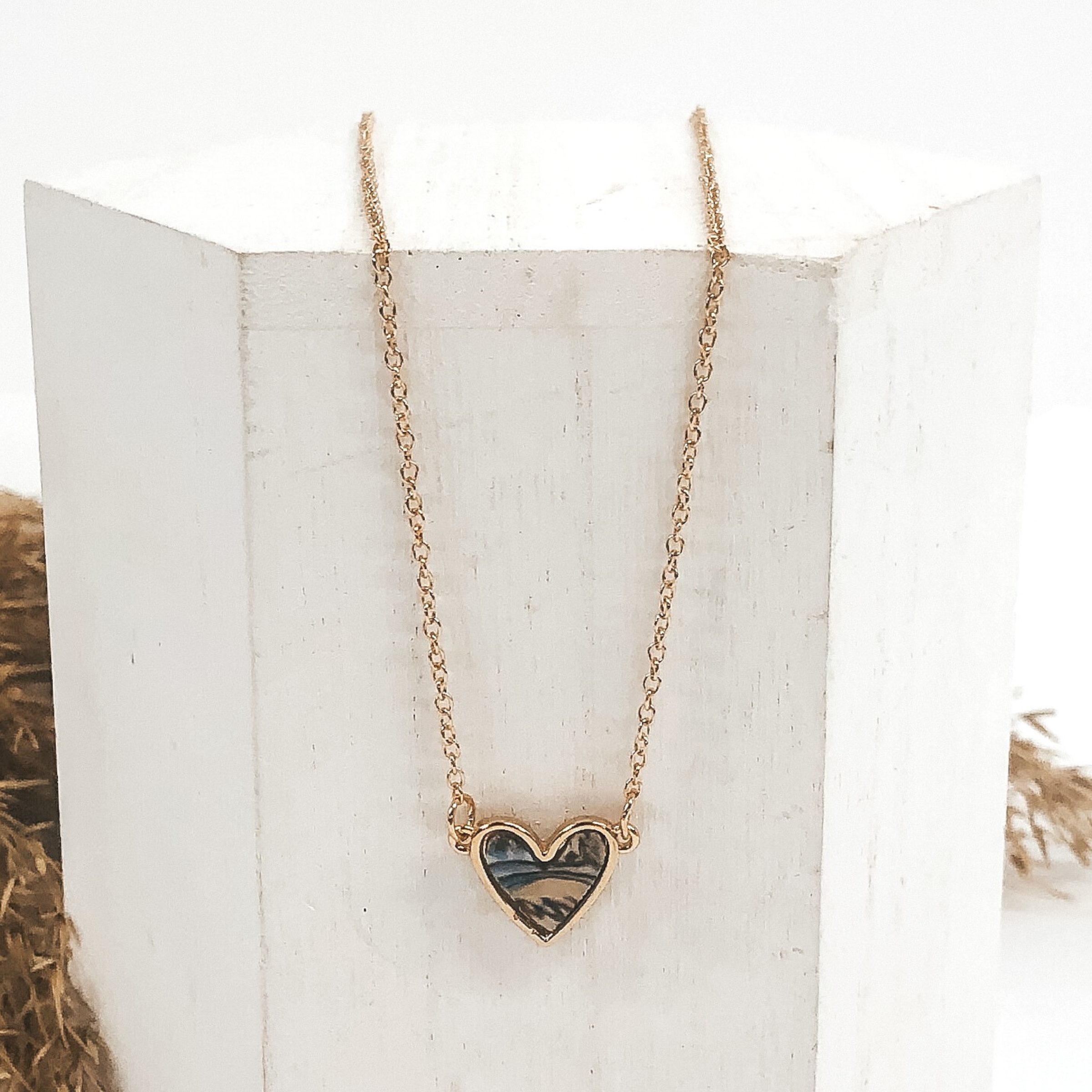 Thin gold chained necklace is a small, abalone shell heart pendant that is outlined in gold. This necklace is pictured laying on a white block on a white background. 