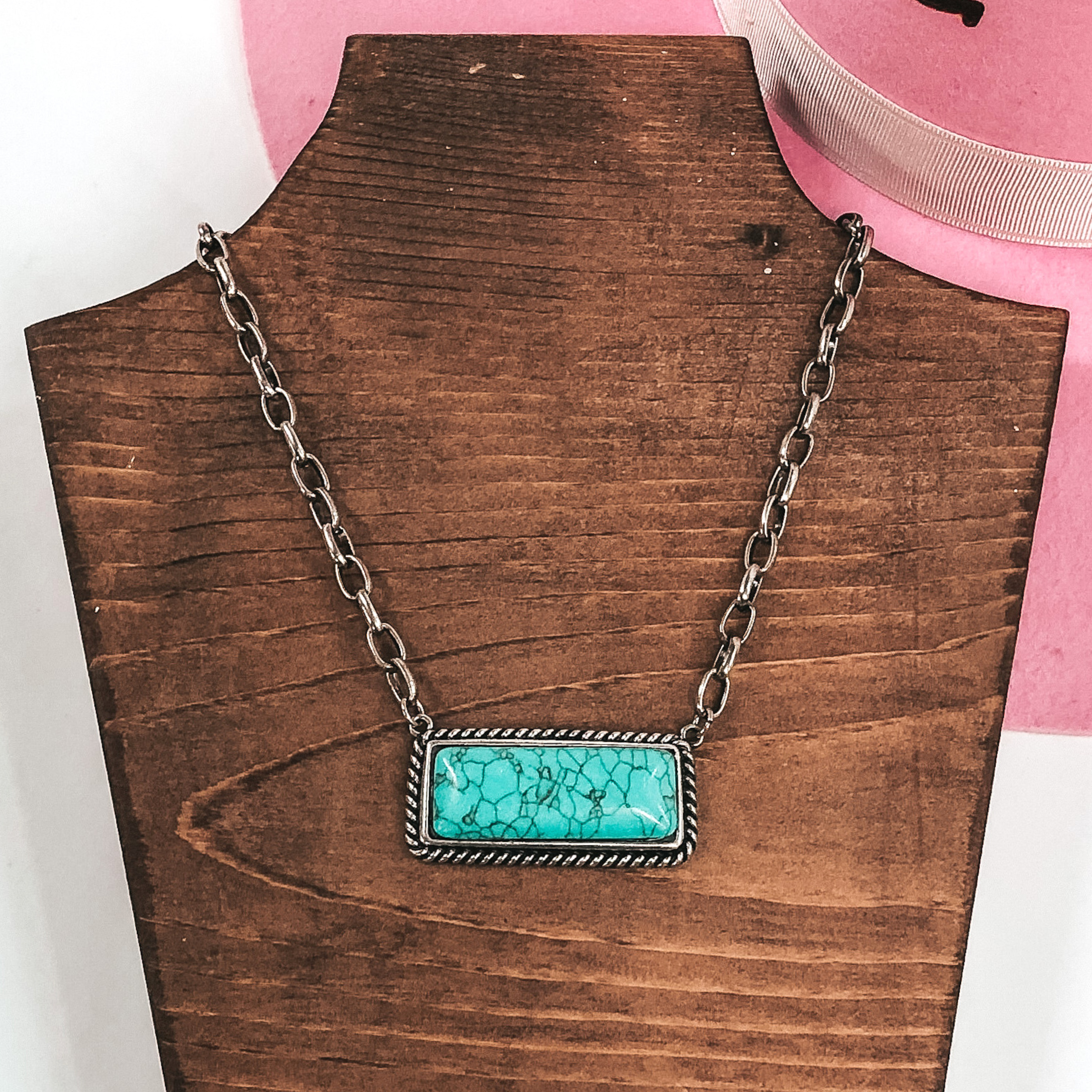 This is a Navajo inspired bar necklace. This necklace includes an oval, silver chained necklace with a bar pendant. The bar pendant is outlined in a silver twist with a center, rectangle stone in turquoise. This necklace is pictured on a dark wood necklace holder on top of a pink hat on a white background. 