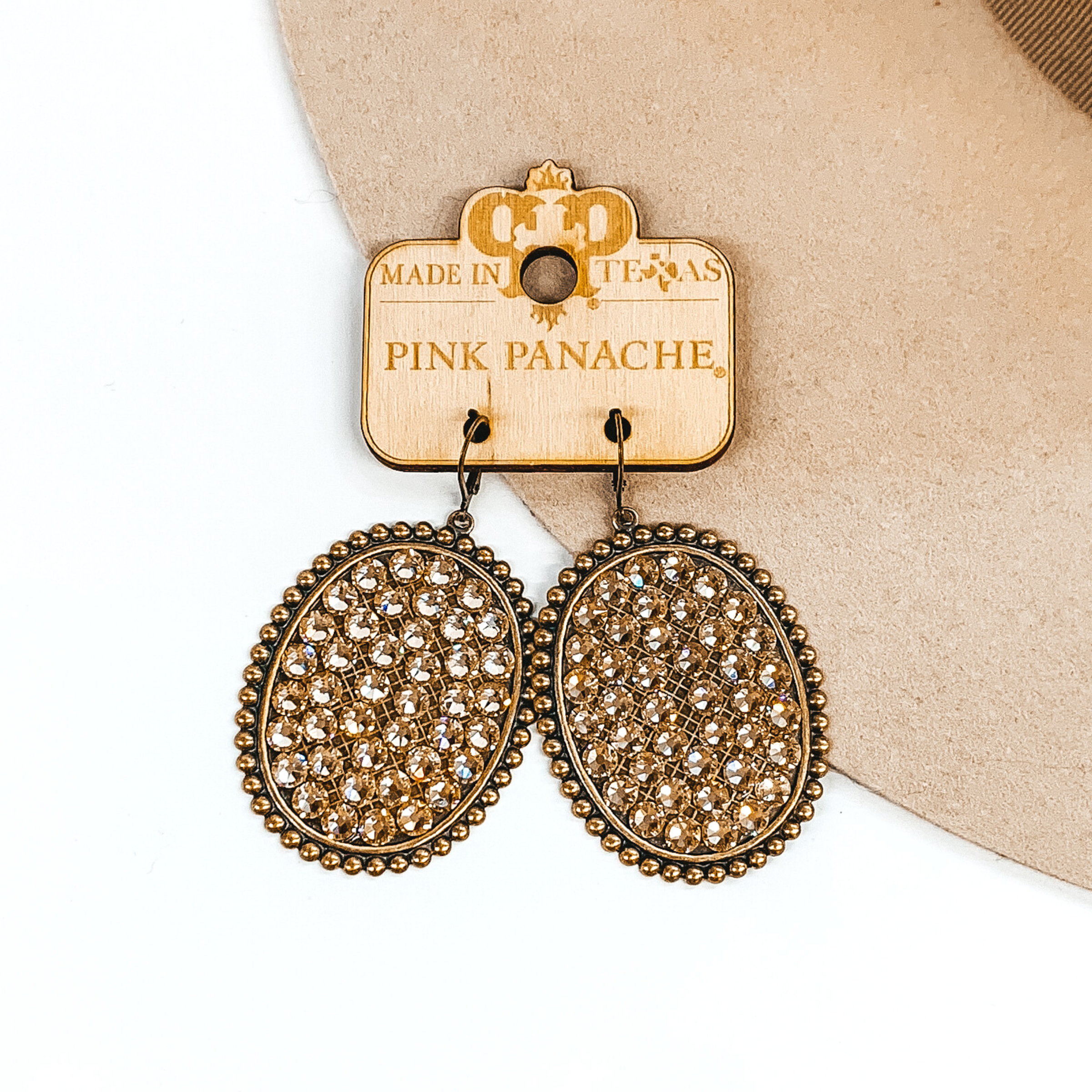 Large, bronze oval dangle earrings. The ovals are outline with bronze circles and has light topaz crystals on the inside of the ovals. These earrings are pictured on a wood earring holder and on a beige and white background.
