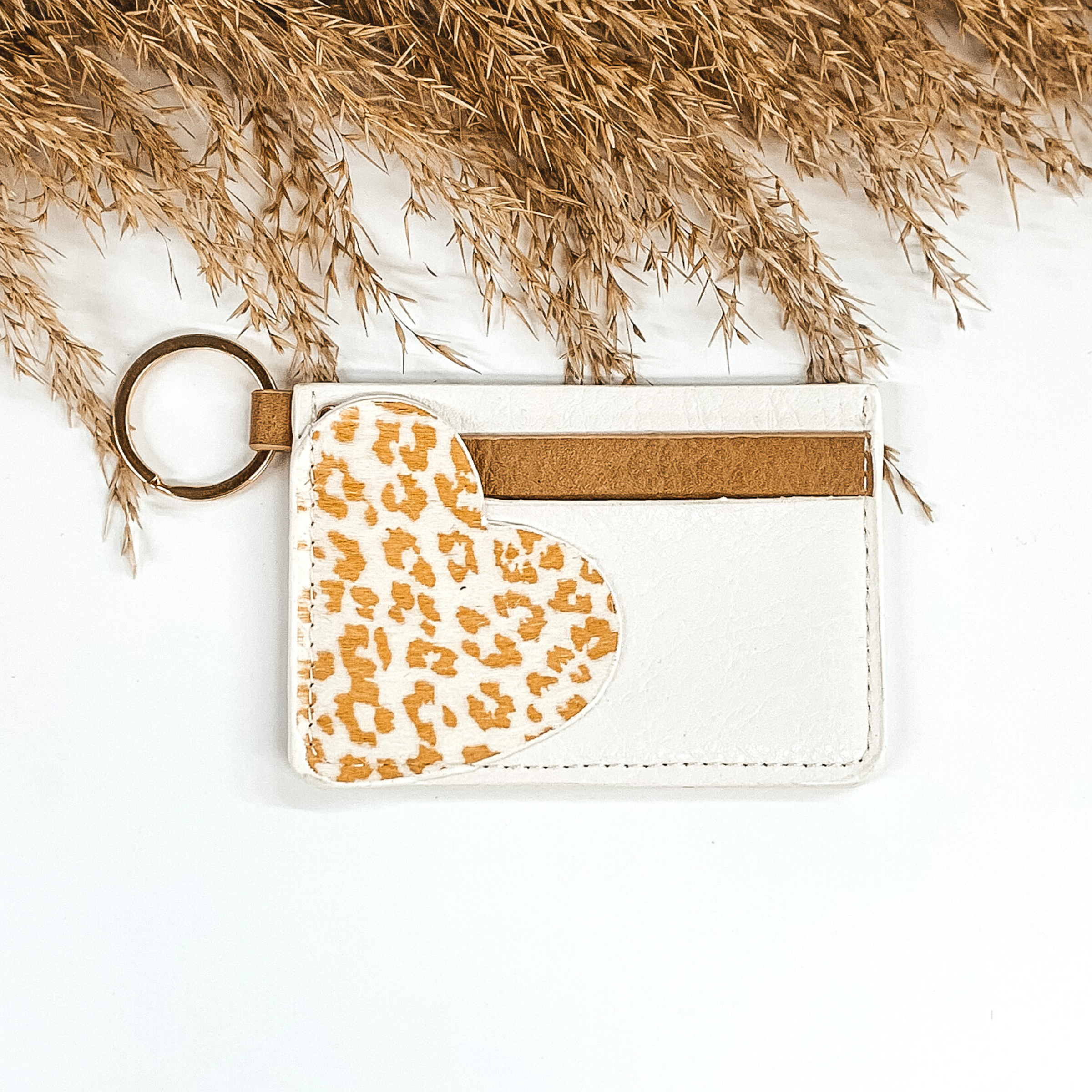 This is a rectangle id key holder that has a gold key ring. On the front you have ivory and tan layer and then a white heart with a tan leopard print is also on the front. It is pictured on a a white background that has brown floral at the top of the picture.