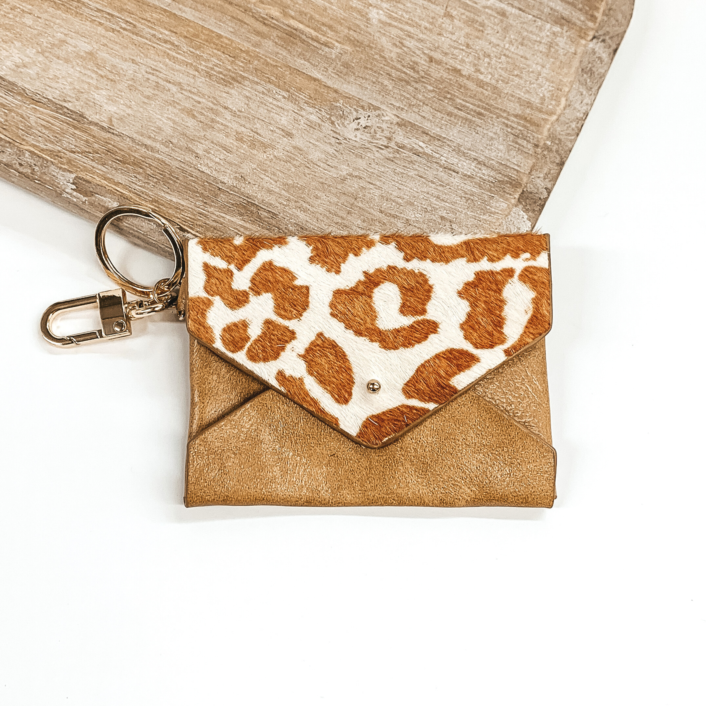 This is a tan wallet with a flap that has a white and tan leopard print. This wallet has a gold key chain. This wallet keychain is pictured laying on a light colored piece of wood on a white background. 