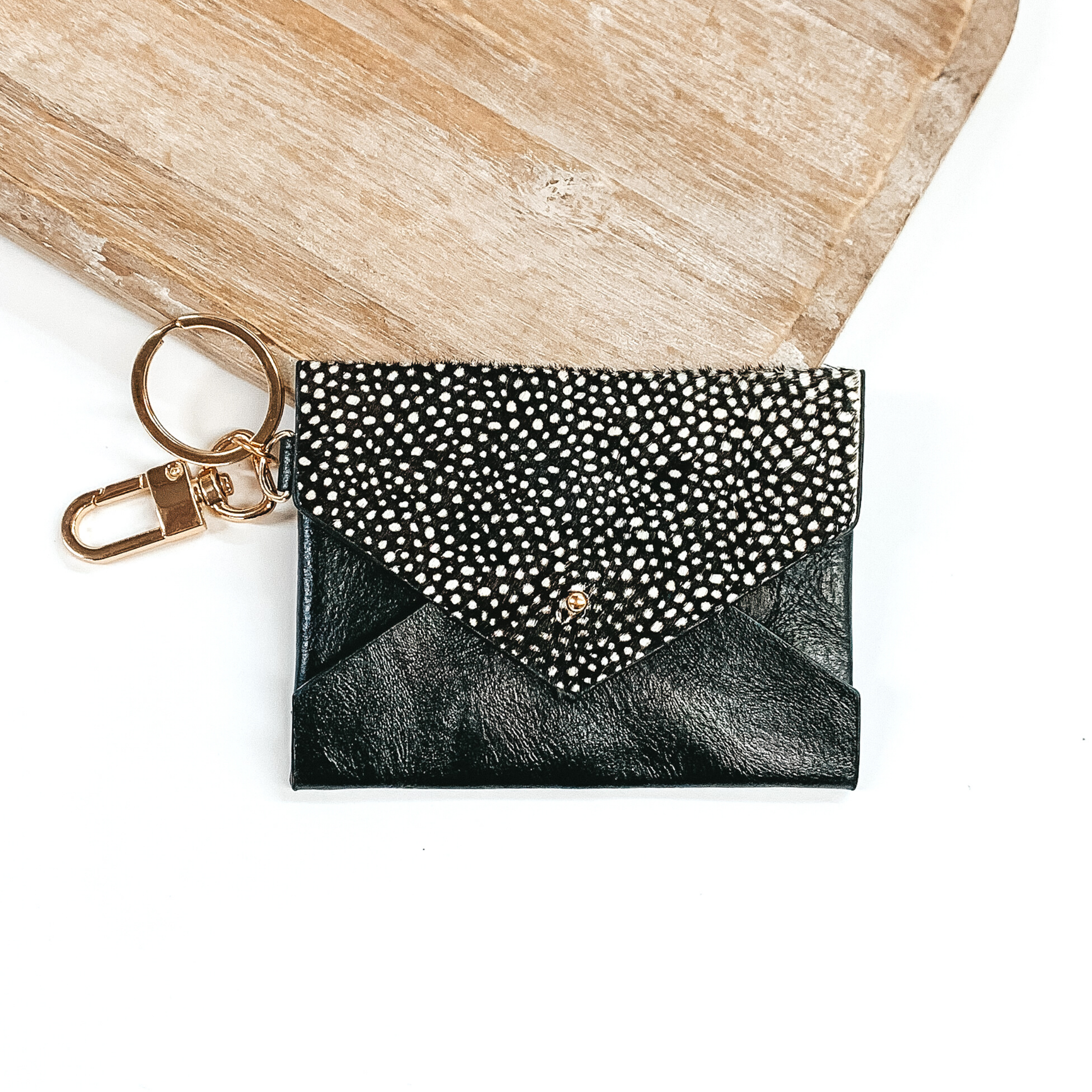 This is a black wallet with a flap that has a black and white dotted print. This wallet has a gold key chain. This wallet keychain is pictured laying on a light colored piece of wood on a white background.