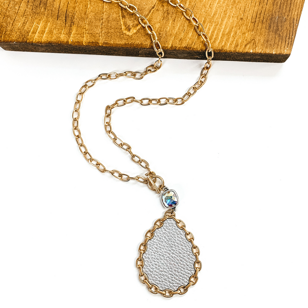 Gold chained necklace with front toggle clasp. This necklace includes a hanging AB cushion cut crystal with a silver back hanging from the toggle clasp. Hanging from the crystal was a teardrop pendant in silver with a gold chained outline. This necklace is pictured partially on a piece of dark wood and on a white background. 