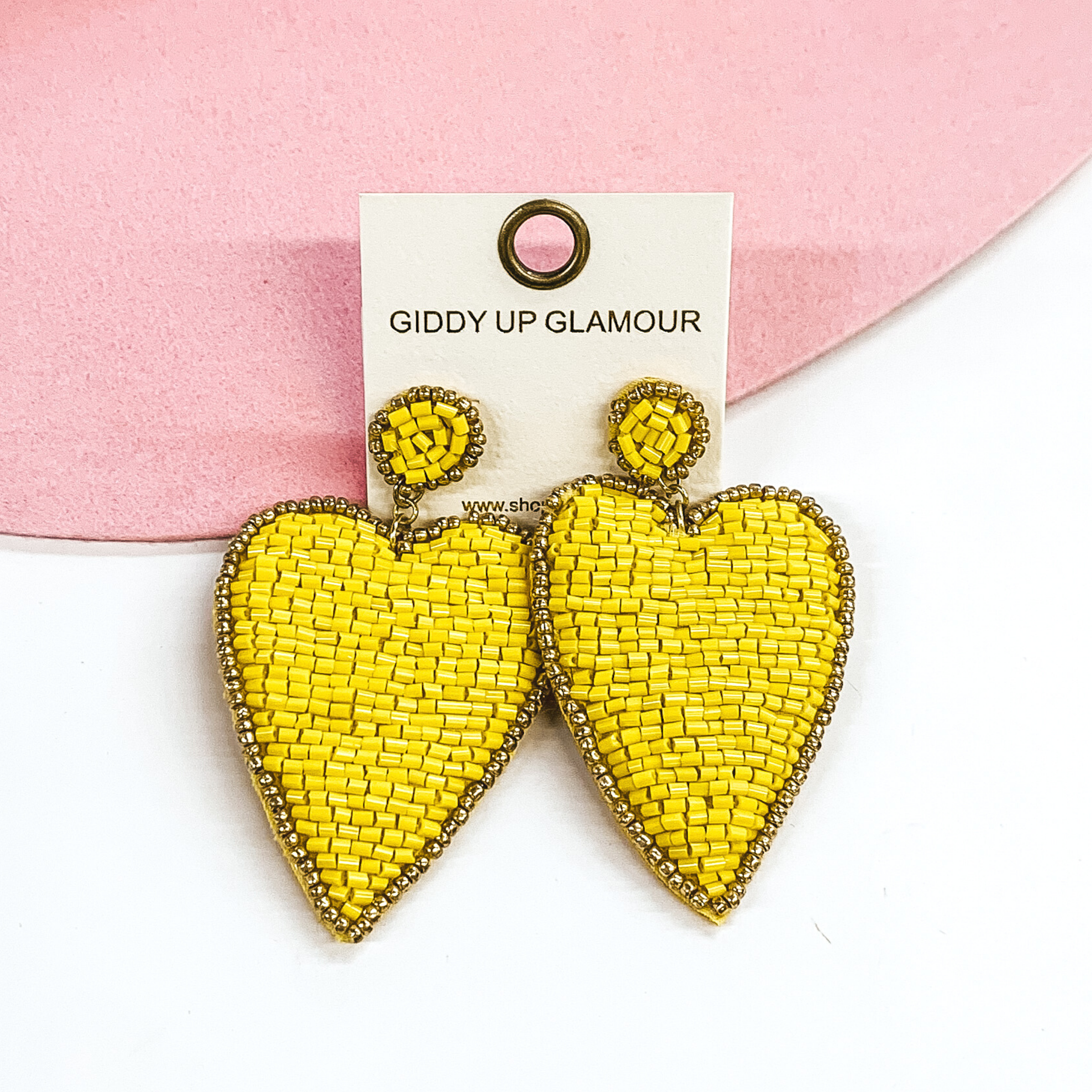 Small circle post studs with a dangle heart. These earrings are covered in yellow beads with a gold outline. These earrings are pictured on a white and pink background. 