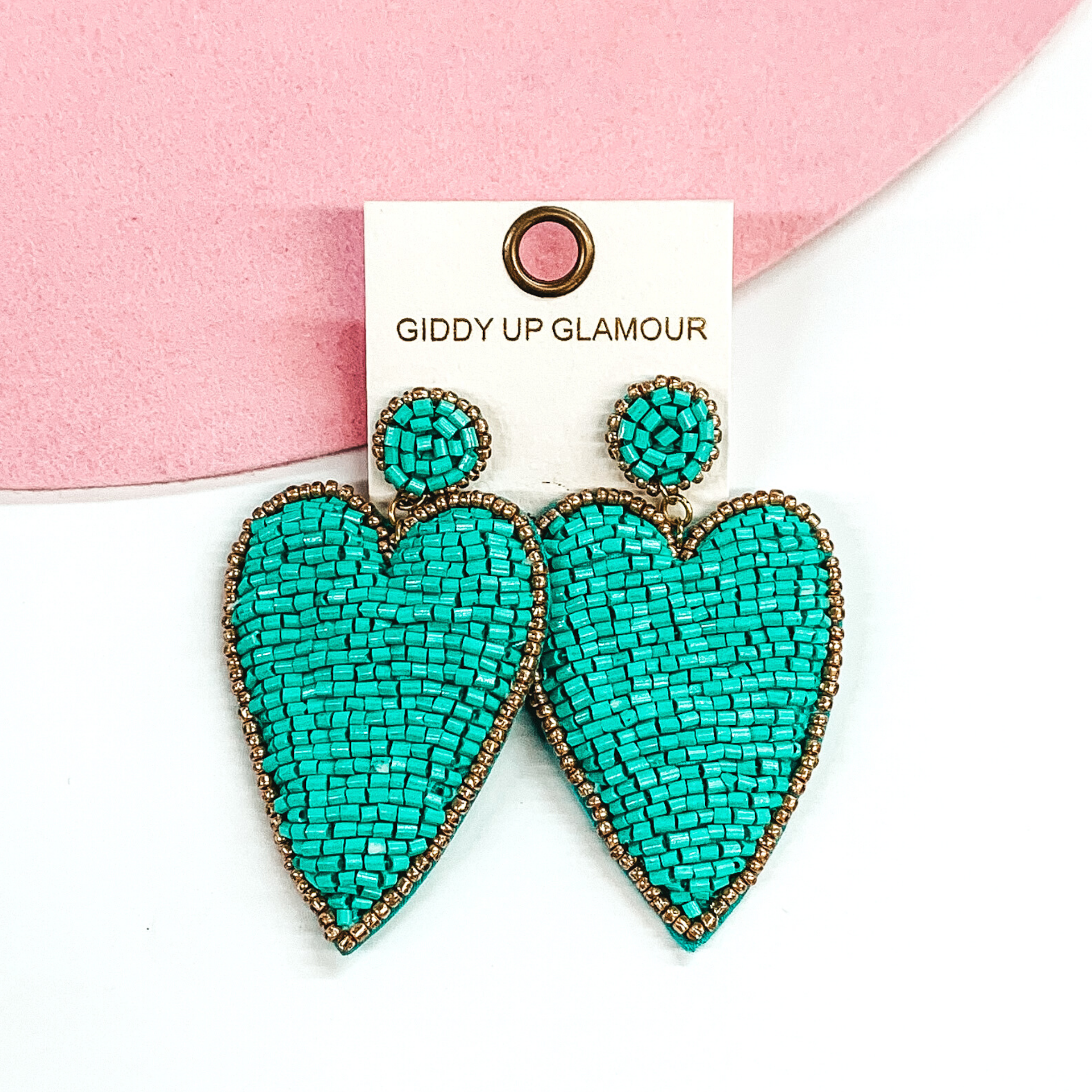Small circle post studs with a dangle heart. These earrings are covered in turquoise beads with a gold outline. These earrings are pictured on a white and pink background.
