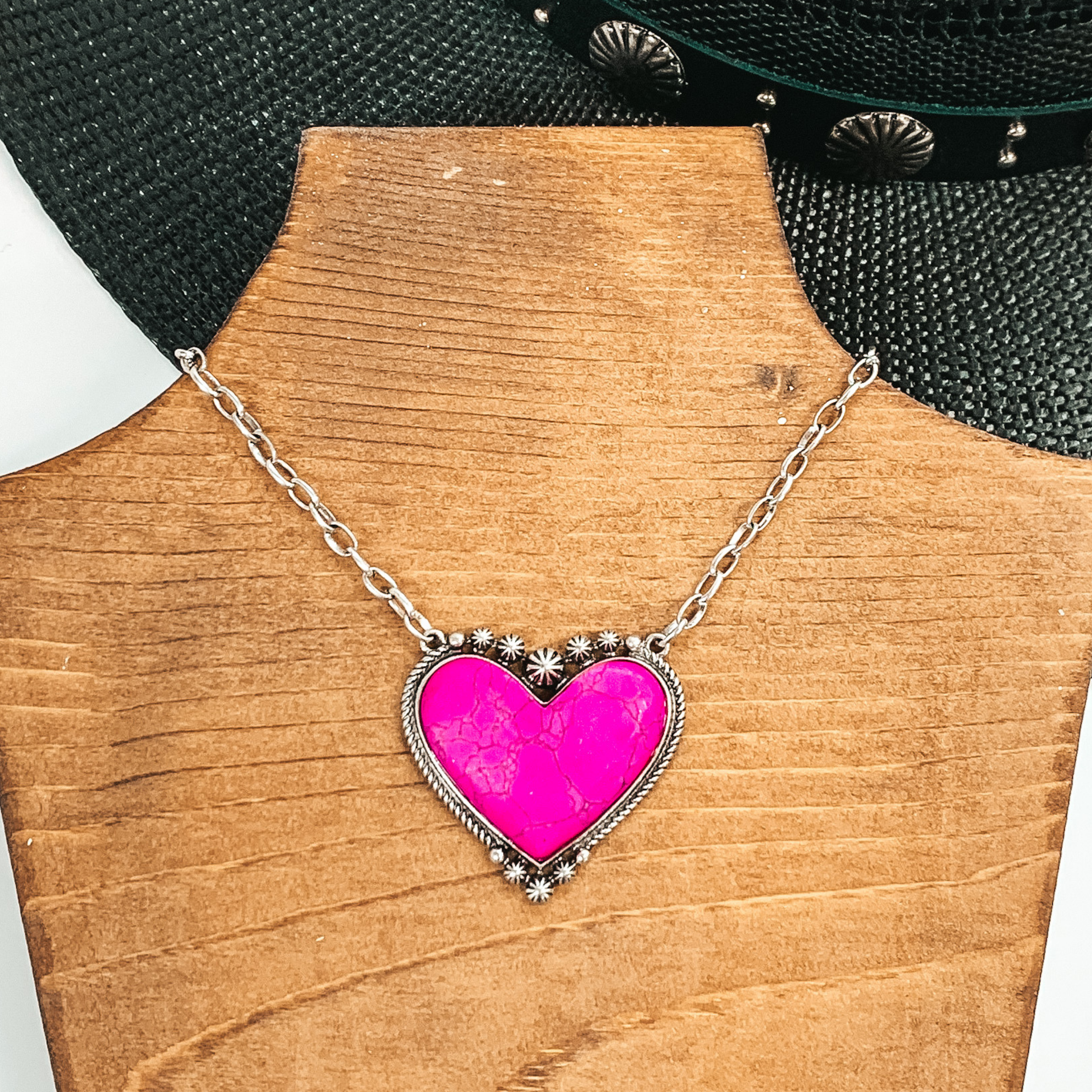 Silver chained necklace with a large pink, heart shaped stone pendant. This necklace is pictured laying on a brown necklace holder on a white and black background.