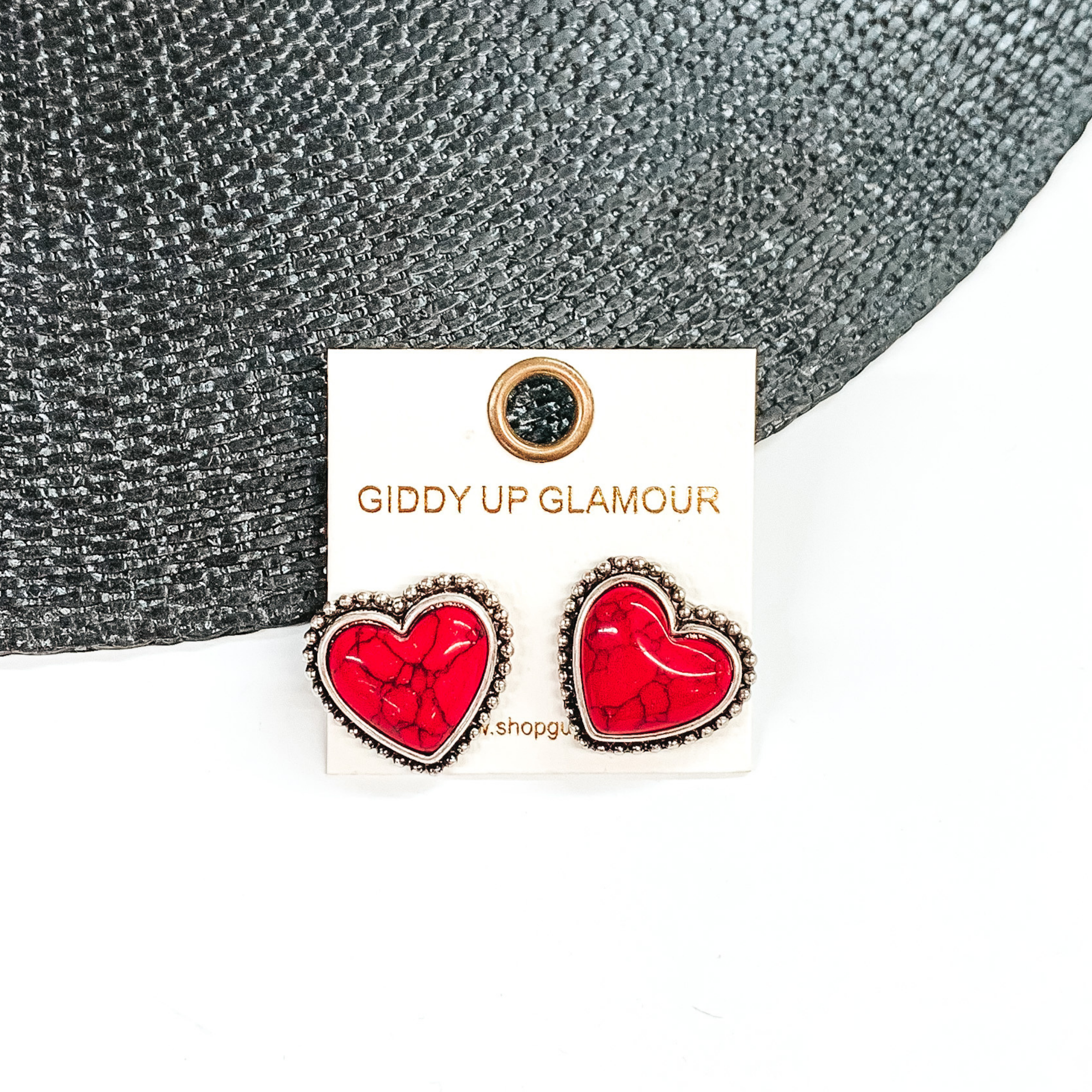Red, heart stone earrings with a silver backing and outline. These earrings are pictured on a white earrings holder. Then pictured on a white and black background.