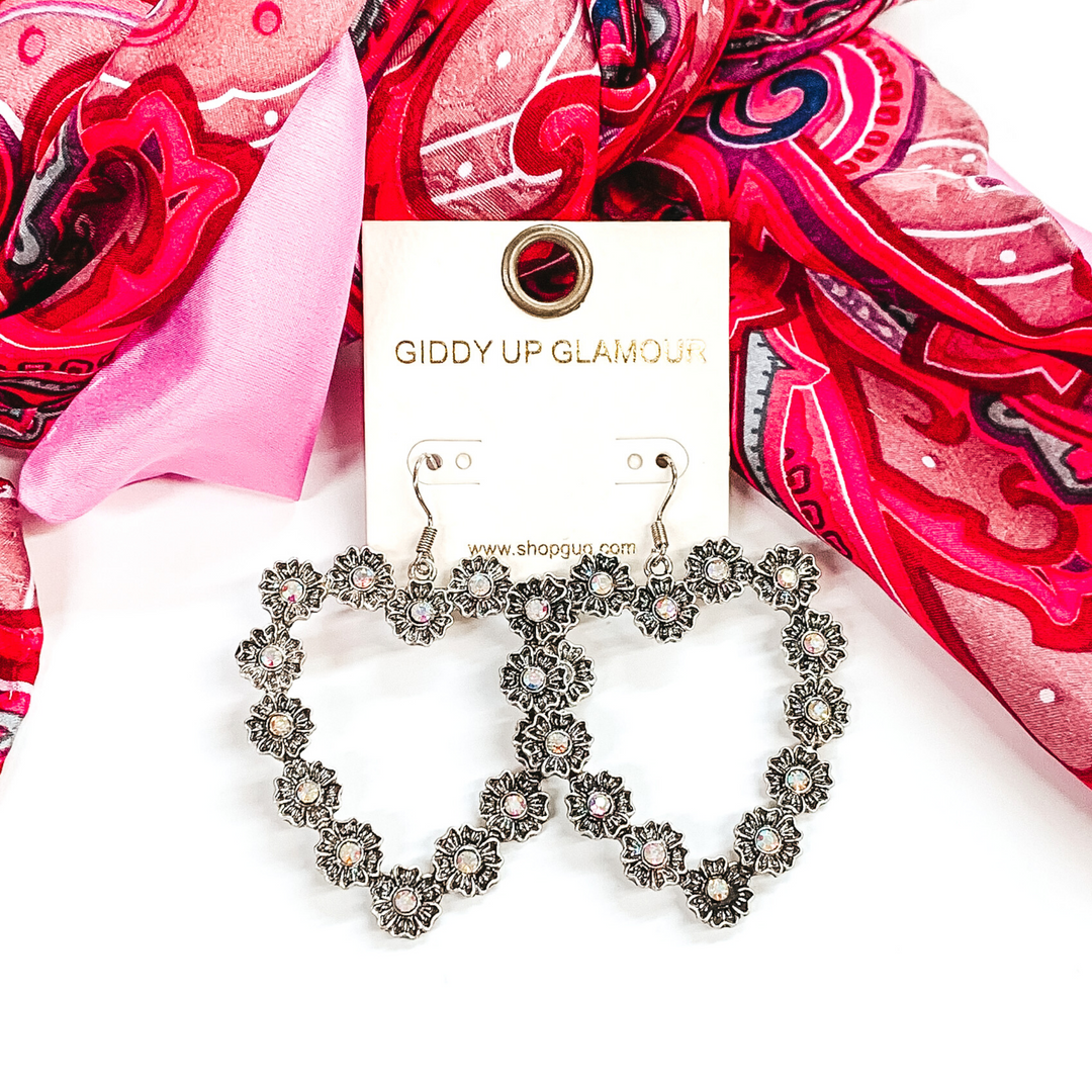 Silver, open heart outline earrings. These earrings have tiny, engraved flowers that make out the shape of the heart. The middle of each flower had a AB crystal. These earrings are pictured layingin front of some pink hued fabric on a white background.