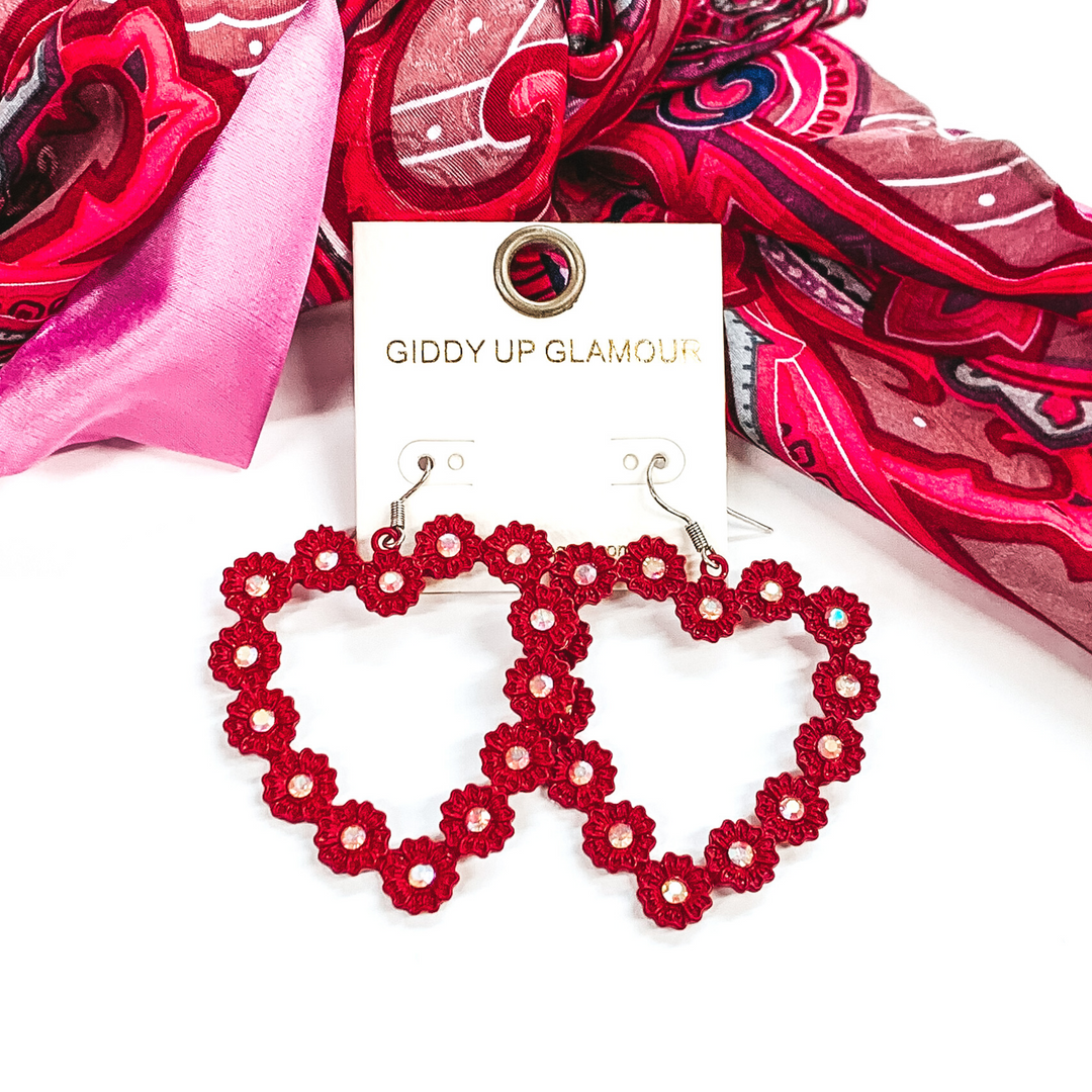 Red, open heart outline earrings. These earrings have tiny, engraved flowers that make out the shape of the heart. The middle of each flower had a AB crystal. These earrings are pictured layingin front of some pink hued fabric on a white background.