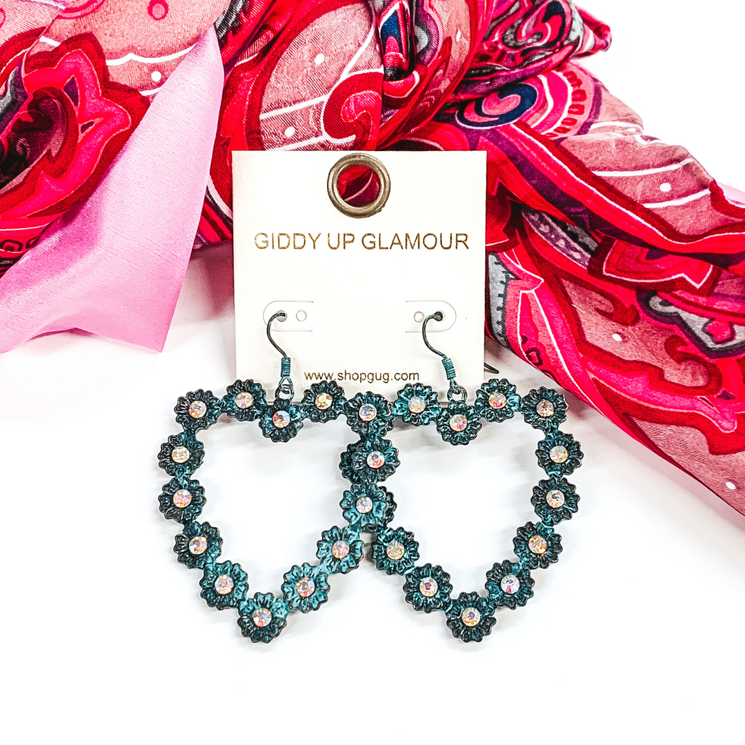 Patina, open heart outline earrings. These earrings have tiny, engraved flowers that make out the shape of the heart. The middle of each flower had a AB crystal. These earrings are pictured layingin front of some pink hued fabric on a white background.