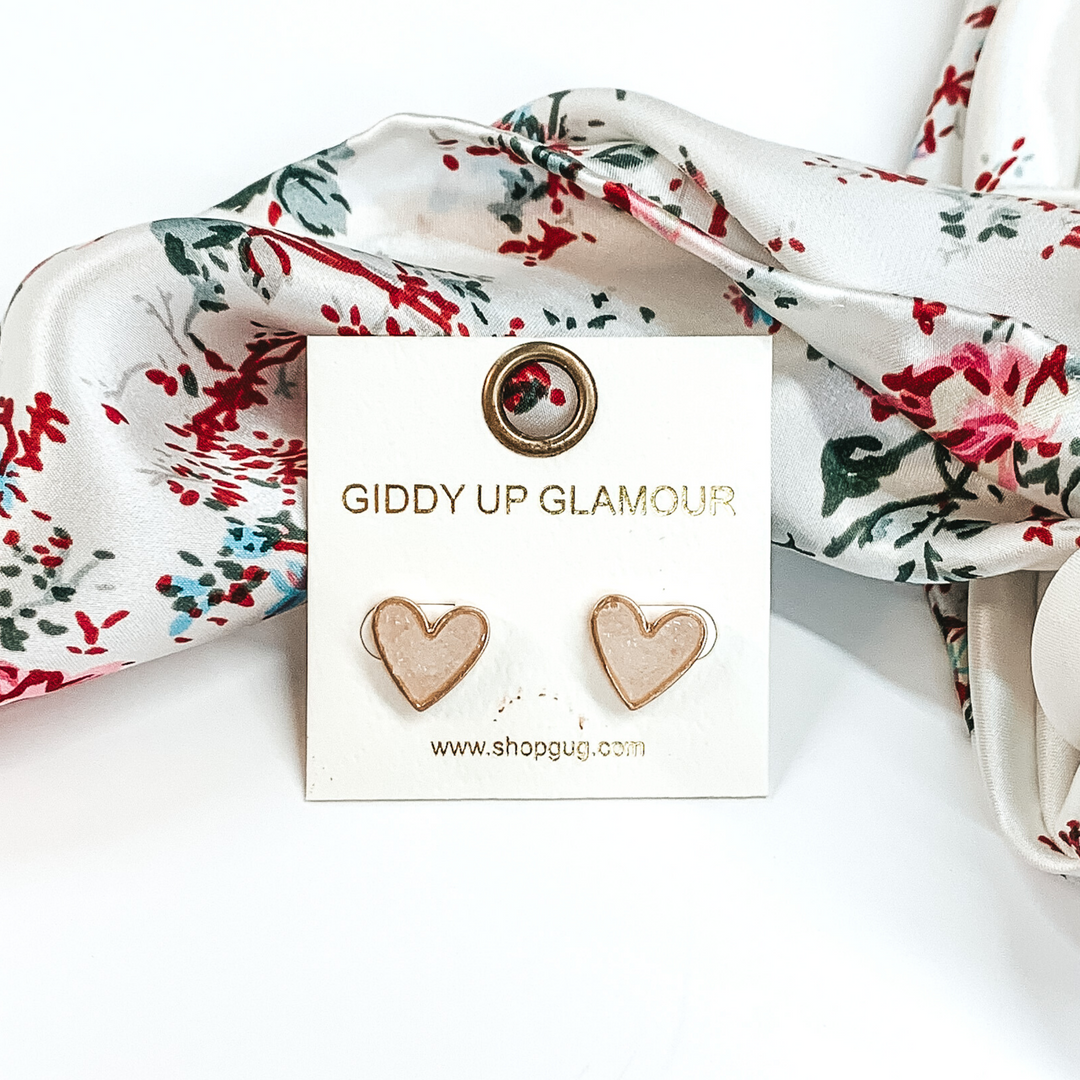 These are heart shaped, white druzy stud earrings pictured on a white earrings holder. These earrings are laying in front of a white wild rag that has colorful florals on it on a white background.