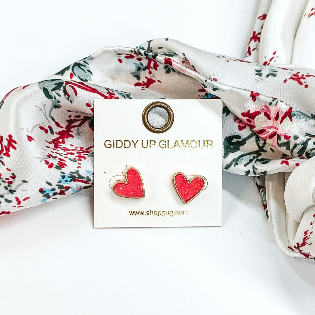 These are heart shaped, pink druzy stud earrings pictured on a white earrings holder. These earrings are laying in front of a white wild rag that has colorful florals on it on a white background.