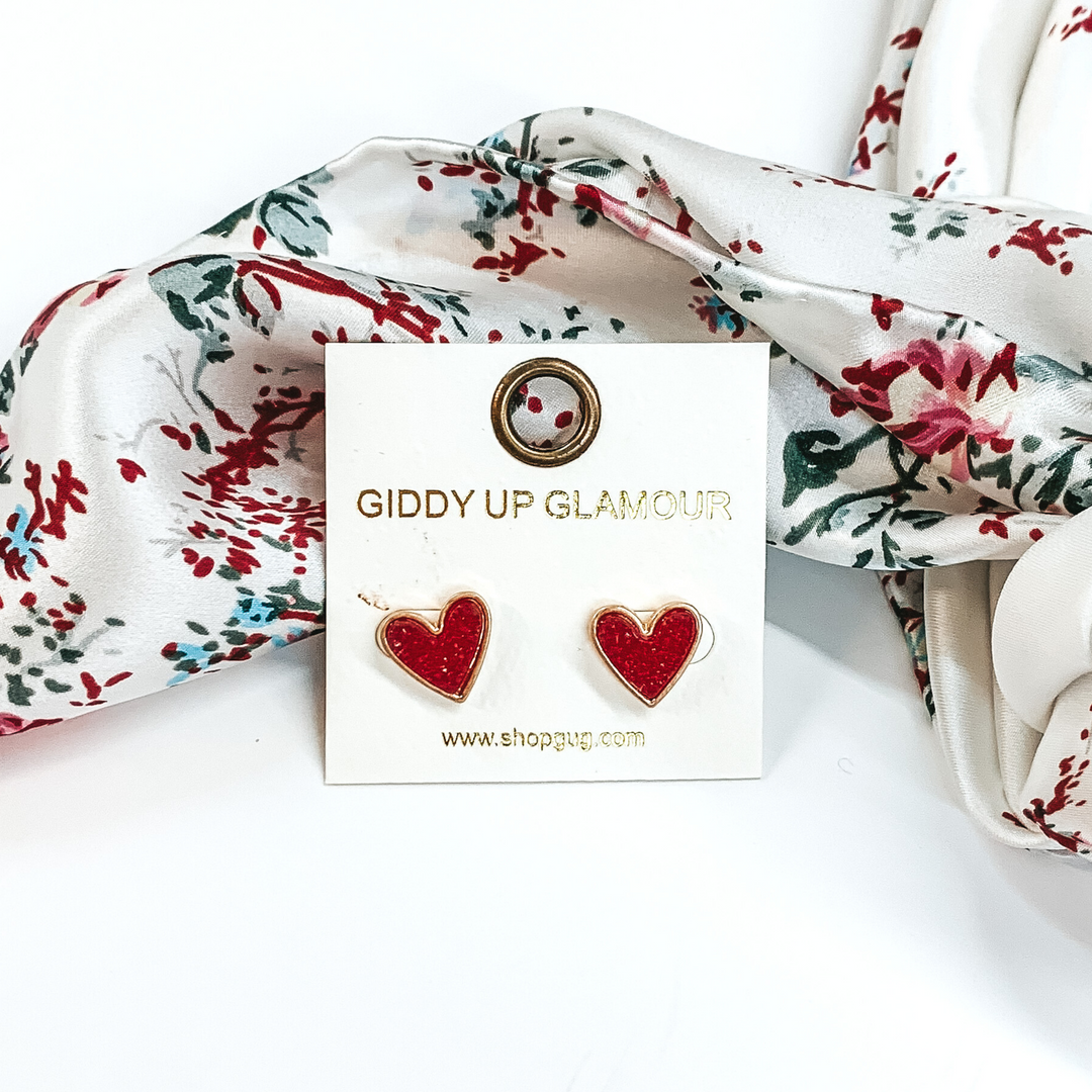 These are heart shaped, maroon druzy stud earrings pictured on a white earrings holder. These earrings are laying in front of a white wild rag that has colorful florals on it on a white background.