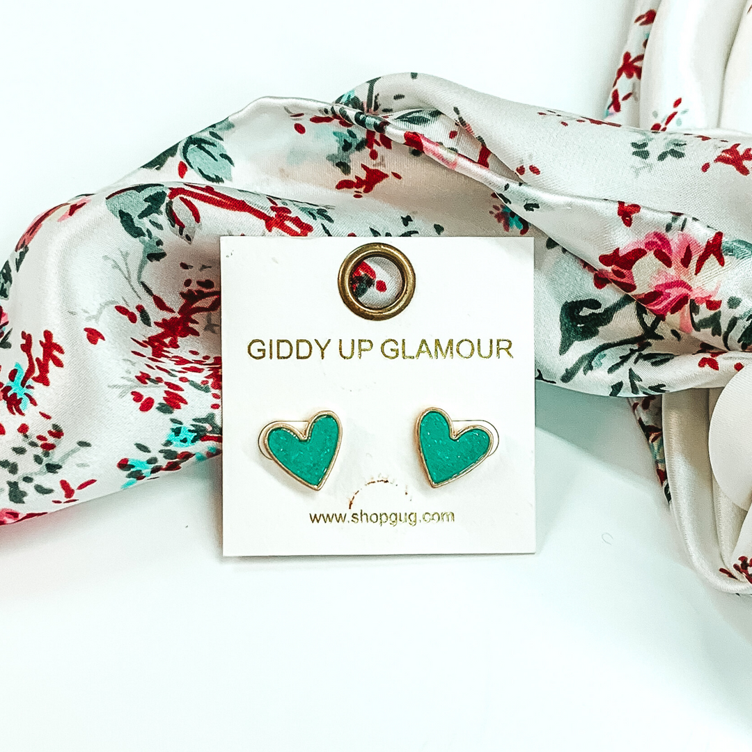 These are heart shaped, mint druzy stud earrings pictured on a white earrings holder. These earrings are laying in front of a white wild rag that has colorful florals on it on a white background.