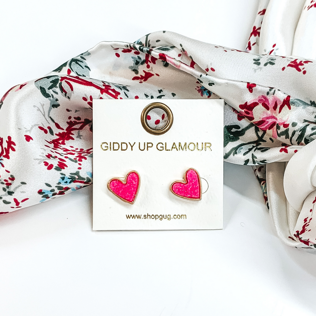 These are heart shaped, black neon pink stud earrings pictured on a white earrings holder. These earrings are laying in front of a white wild rag that has colorful florals on it on a white background.