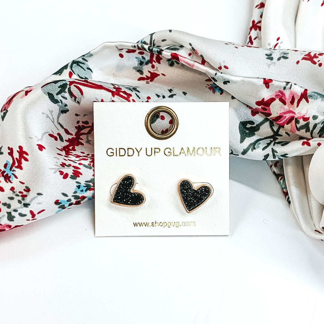 These are heart shaped, black druzy stud earrings pictured on a white earrings holder. These earrings are laying in front of a white wild rag that has colorful florals on it on a white background. 