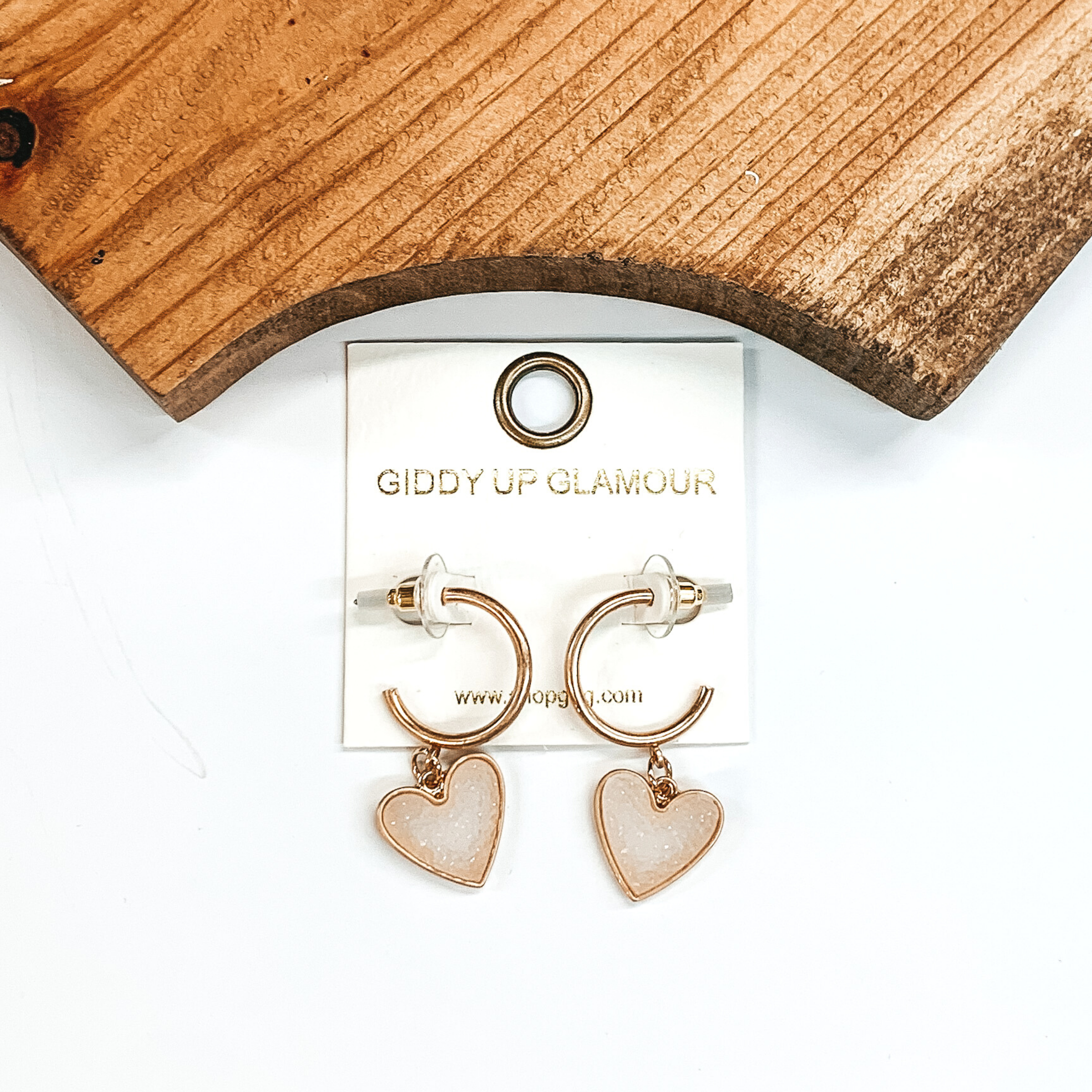 Gold hoop earrings with a white druzy heart charm at the bottom of the hoop. These earring are pictured on a white background with a piece of wood pictured at the top of the picture.