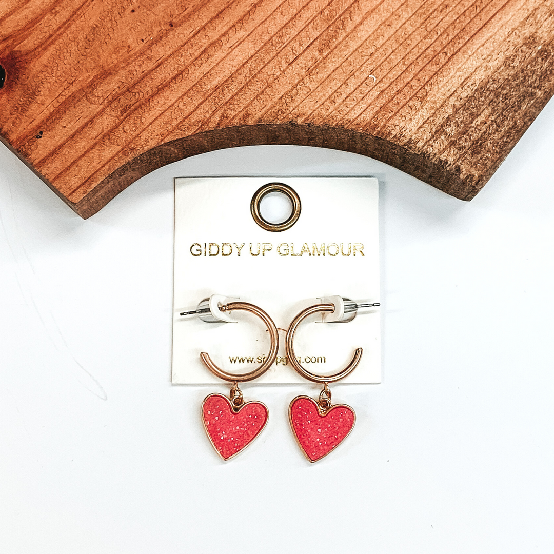 Gold hoop earrings with a pink druzy heart charm at the bottom of the hoop. These earring are pictured on a white background with a piece of wood pictured at the top of the picture.