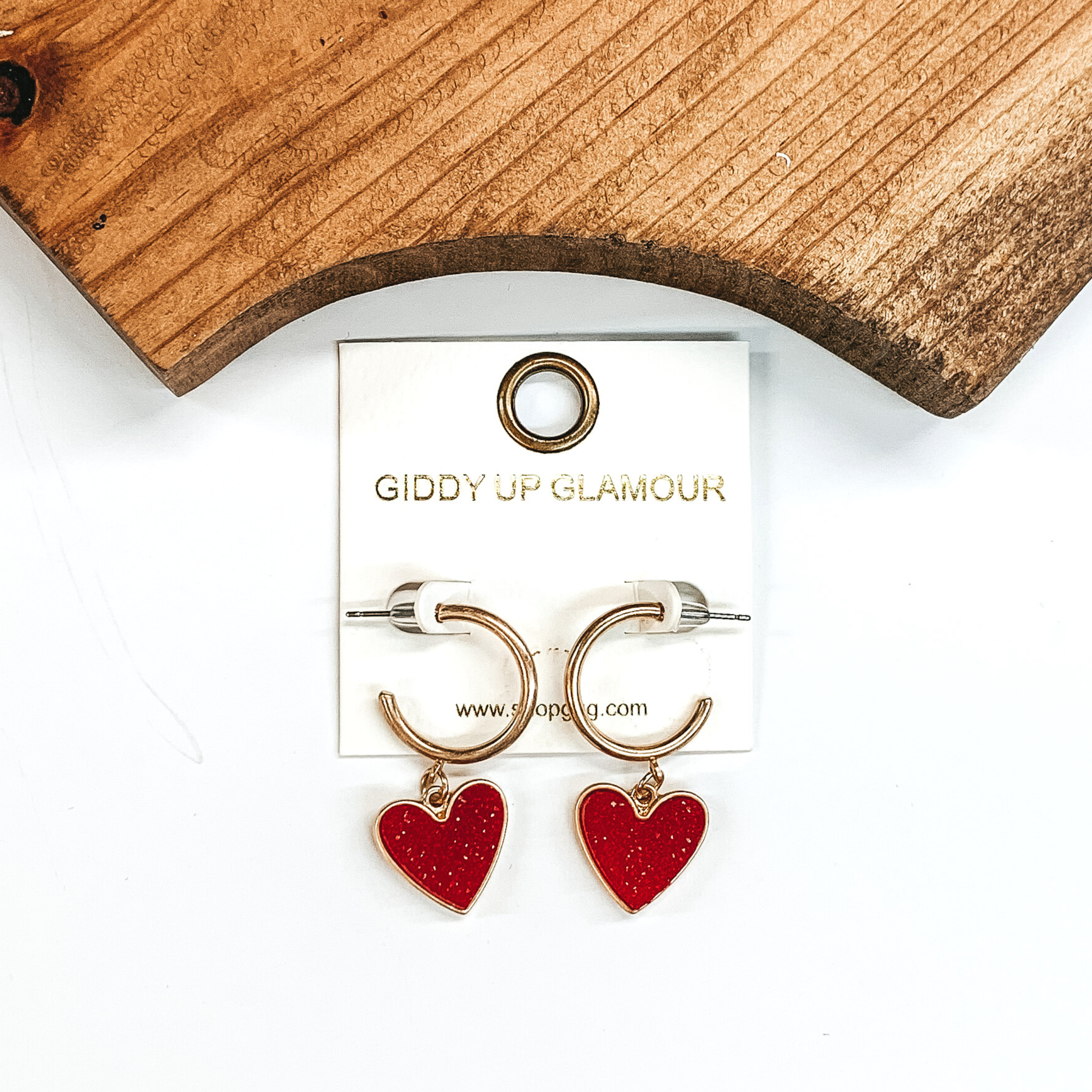 Gold hoop earrings with a maroon druzy heart charm at the bottom of the hoop. These earring are pictured on a white background with a piece of wood pictured at the top of the picture.