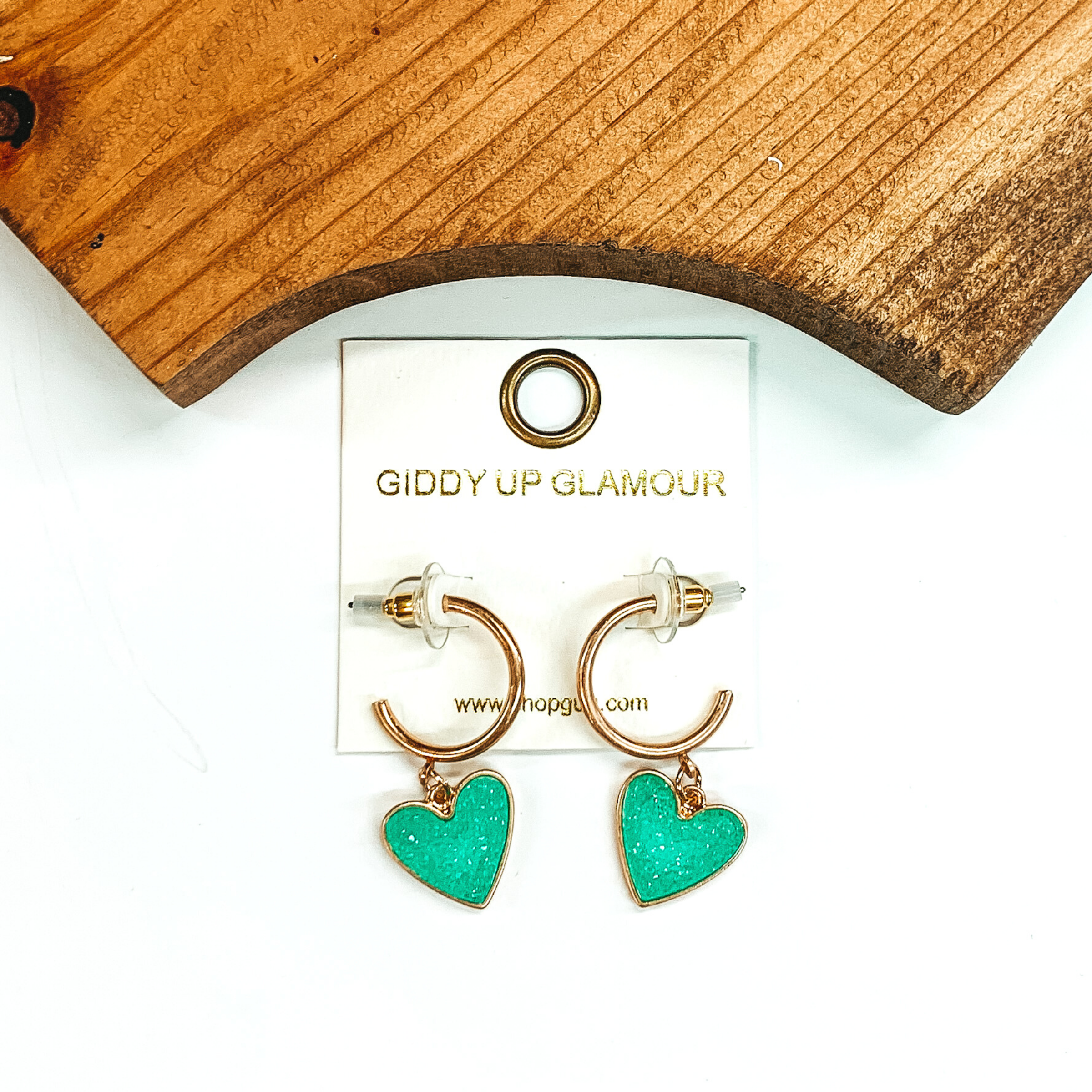 Gold hoop earrings with a mint druzy heart charm at the bottom of the hoop. These earring are pictured on a white background with a piece of wood pictured at the top of the picture.