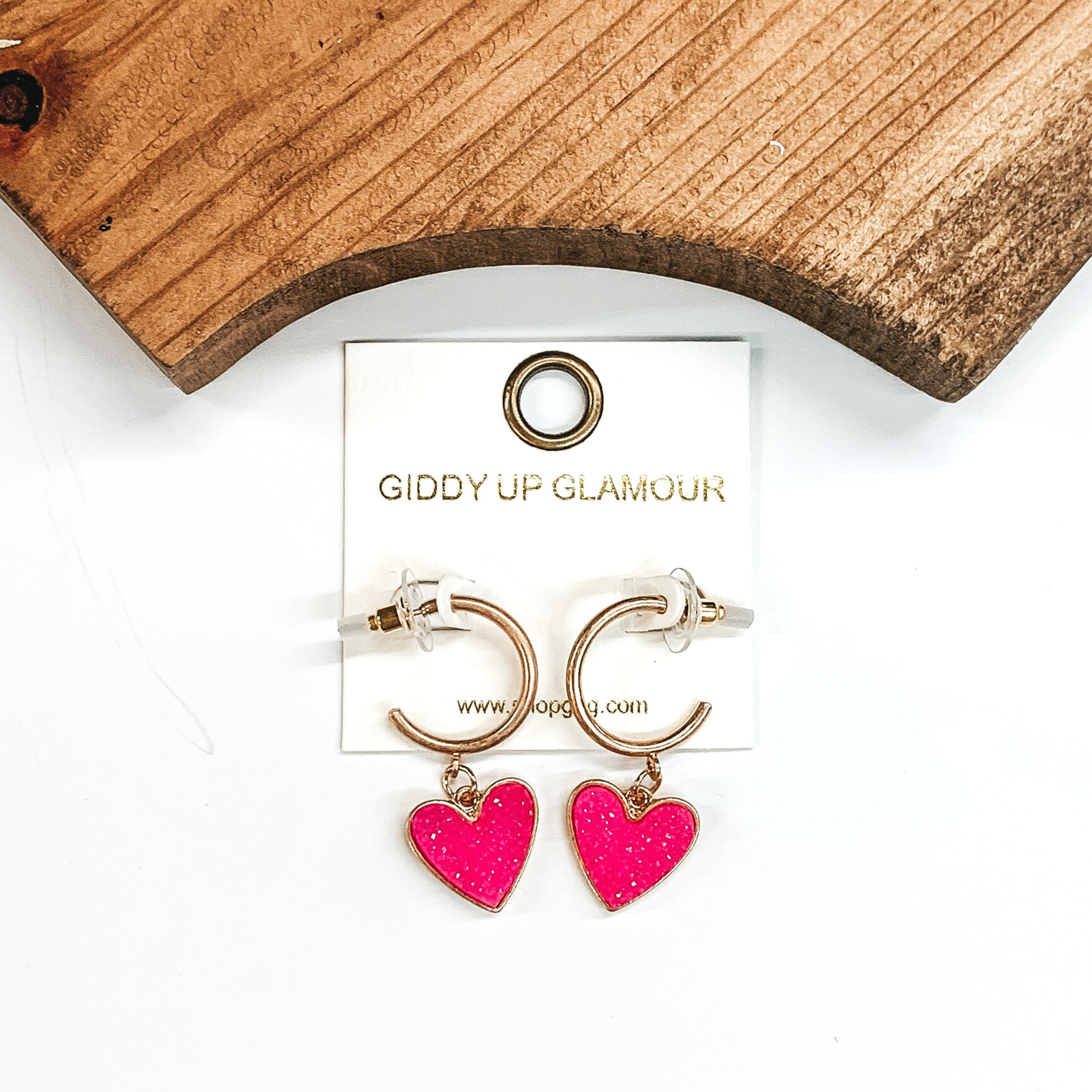 Gold hoop earrings with a neon pink druzy heart charm at the bottom of the hoop. These earring are pictured on a white background with a piece of wood pictured at the top of the picture.
