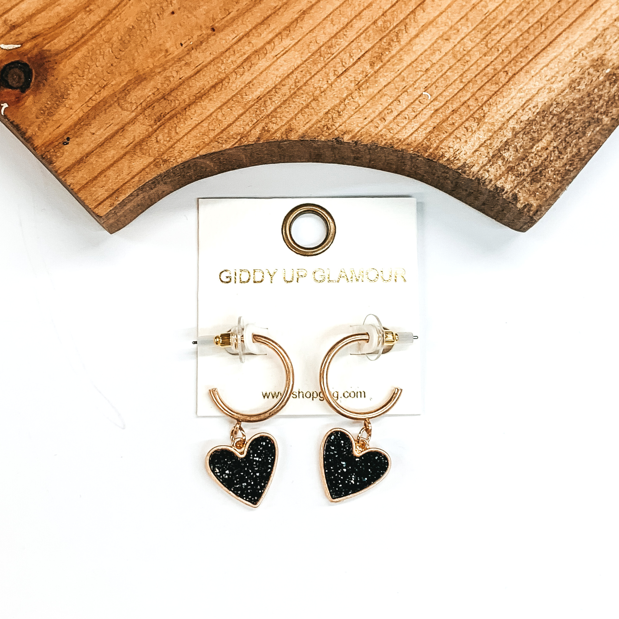 Gold hoop earrings with a black druzy heart charm at the bottom of the hoop. These earring are pictured on a white background with a piece of wood pictured at the top of the picture. 