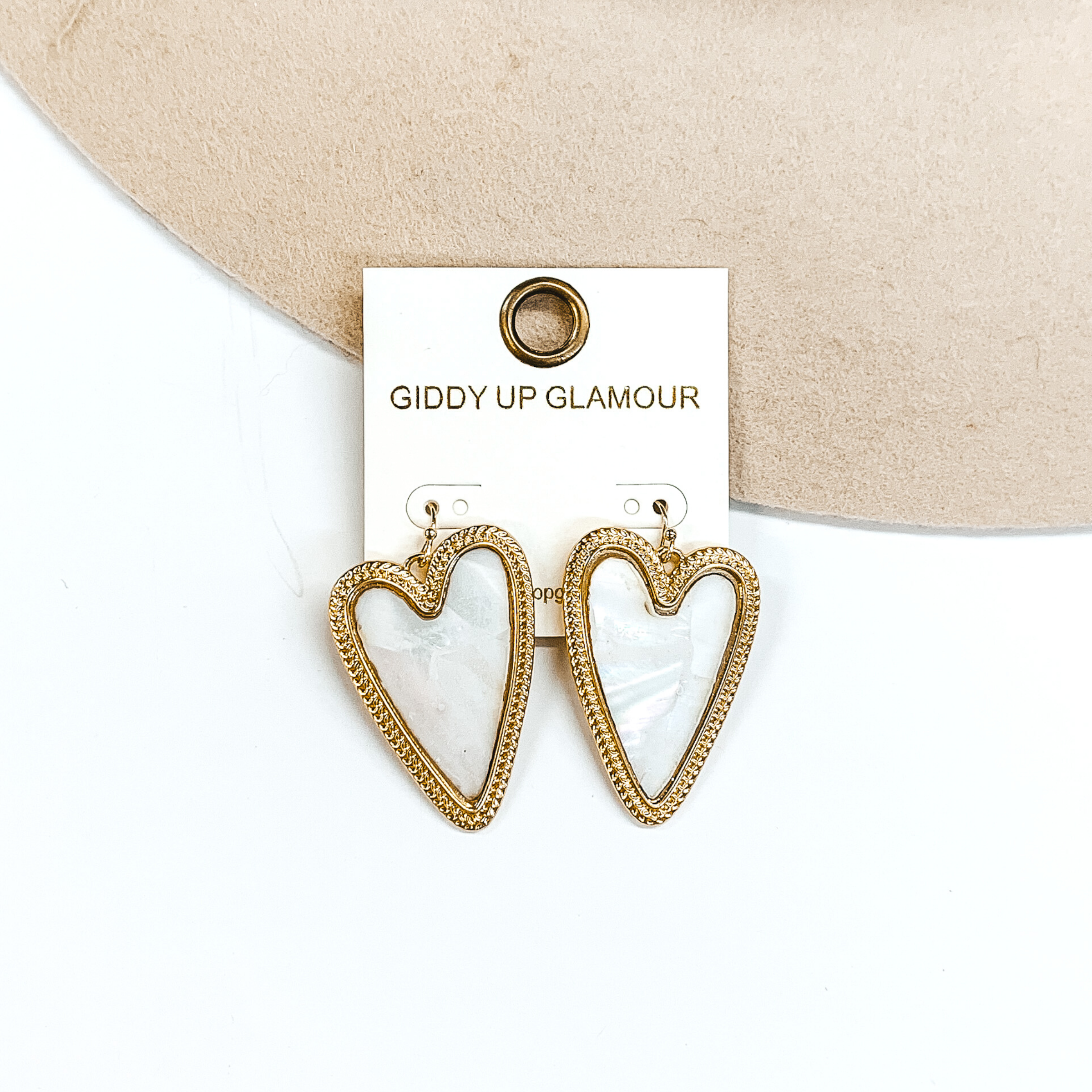 Ivory shell, heart shaped dangle earrings outlined in gold. These earrings are pictured on a white and beige background. 