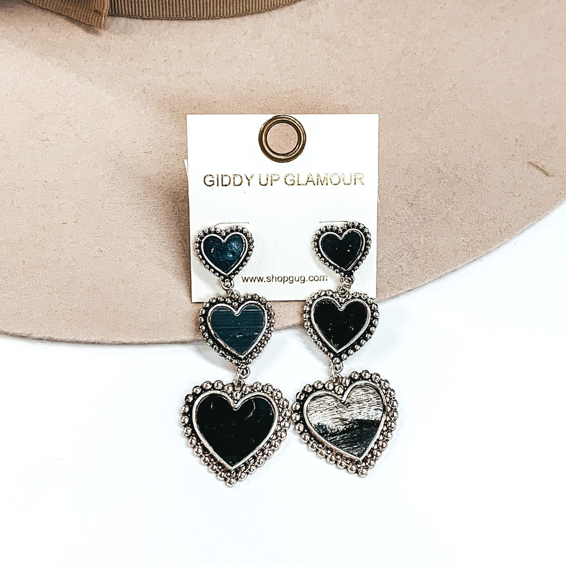 Three tiered heart drop earrings with hearts going from smallest to largest. These earrings have a silver outline with a black shell inlay. These earrings are pictured on a white and beige background.