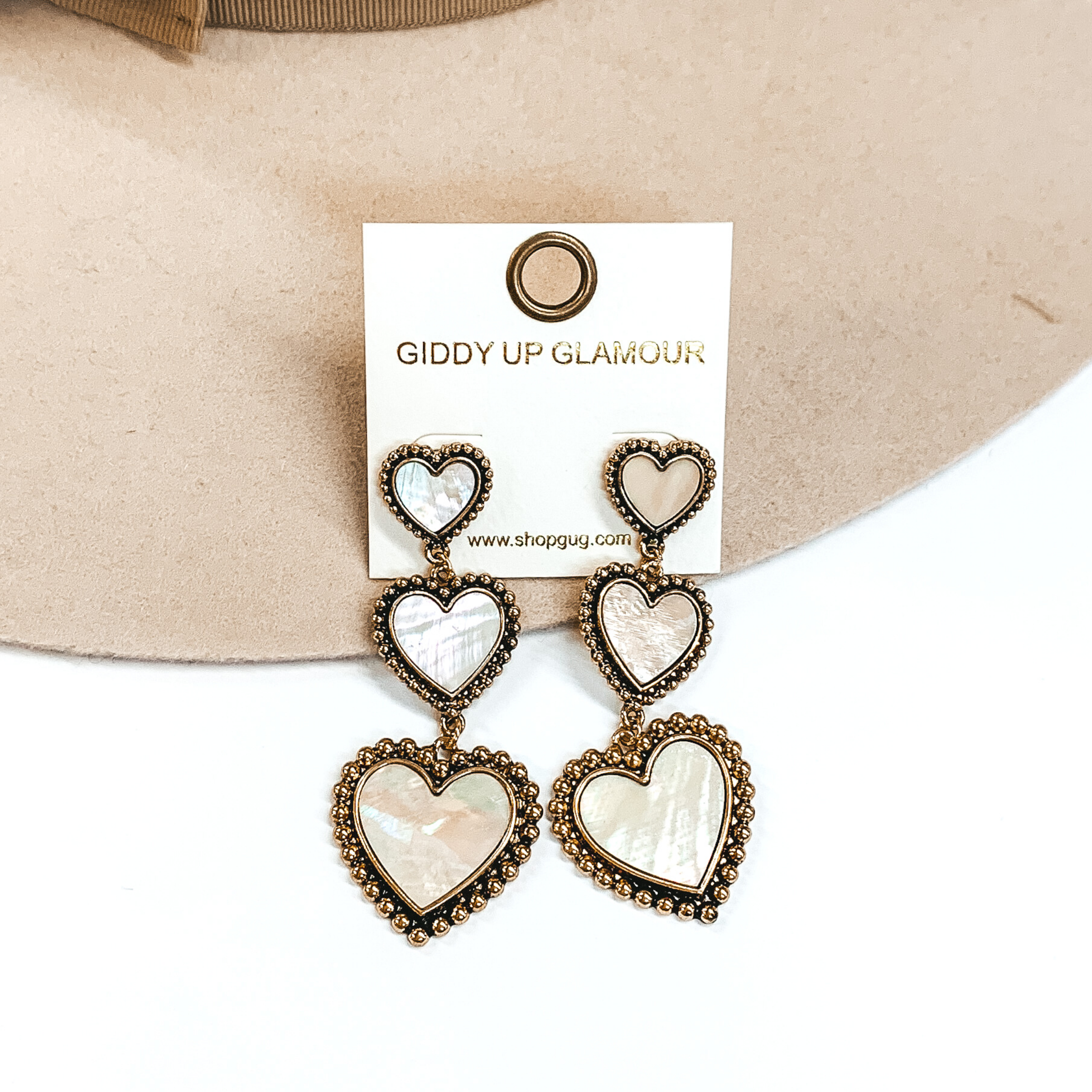 Three tiered heart drop earrings with hearts going from smallest to largest. These earrings have a gold outline with an ivory shell inlay. These earrings are pictured on a white and beige background. 