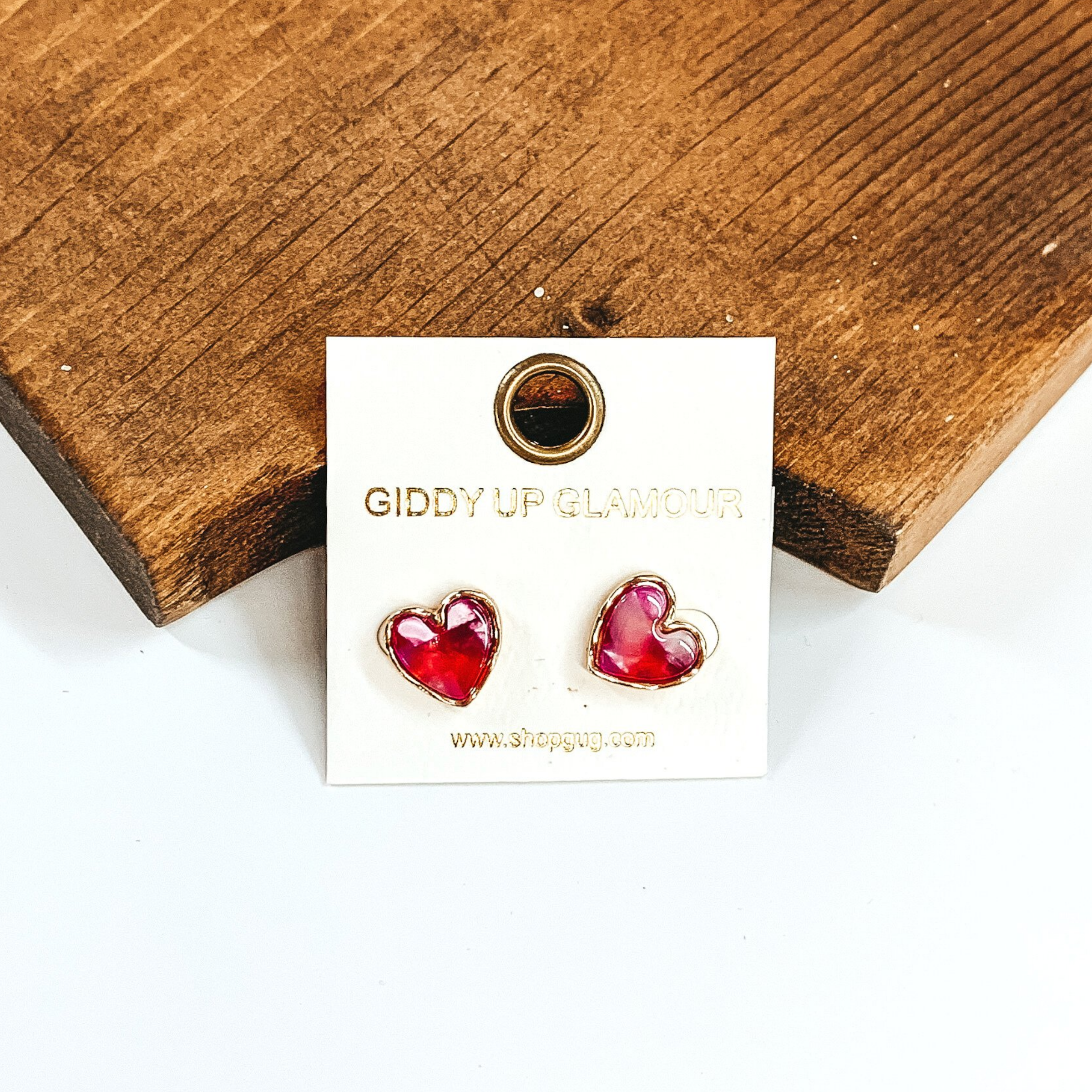 These are heart shaped stud earrings. It has a mix of red, pink, purple, and white on the earrings with a gold outline. These earrings are pictured on a white earrings holder that is laying in front of a piece of wood on a white background.
