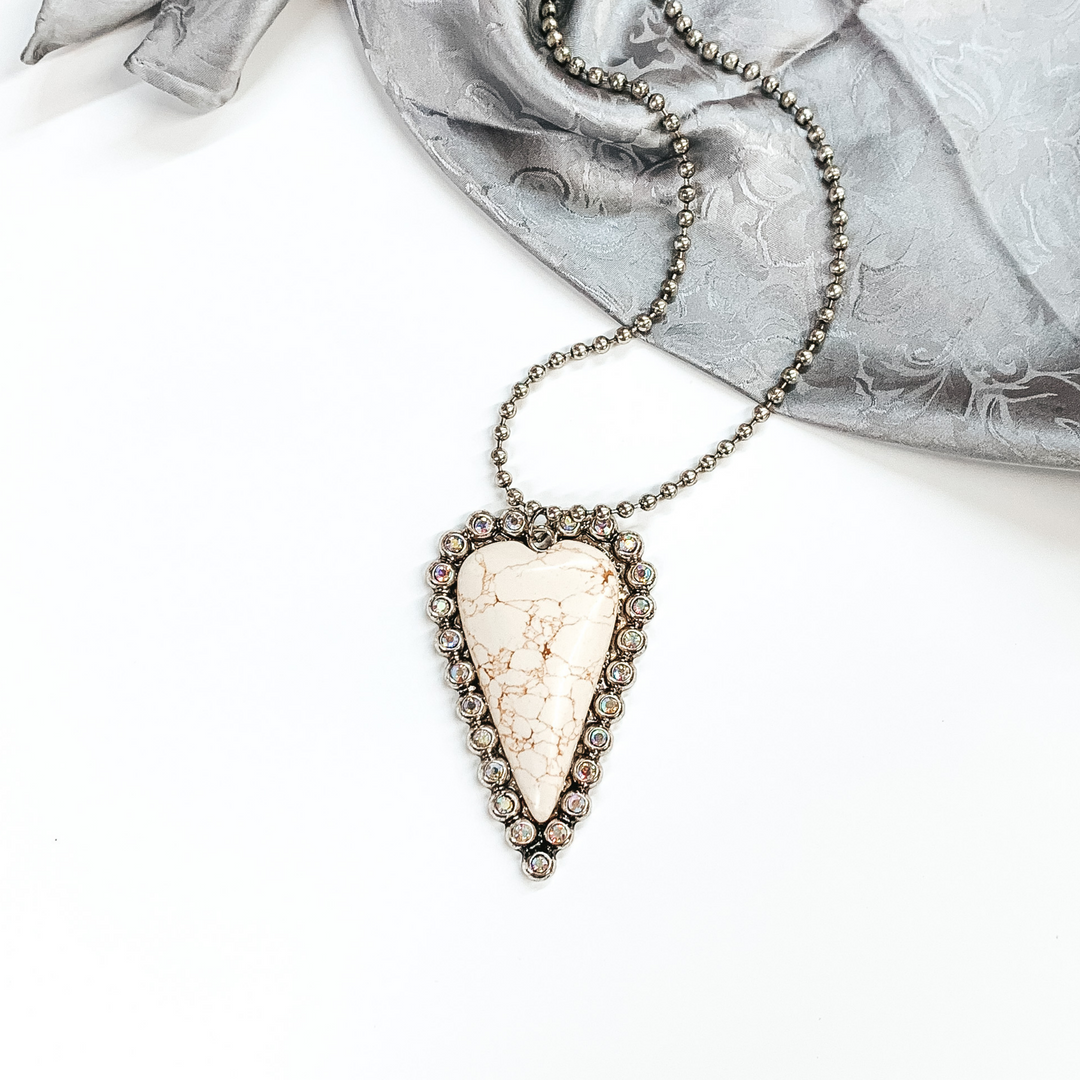 Silver beaded necklace with big heart pendant. The heart pendant is a ivory stone with a silver, ab crystal outline. This necklace is pictured on a white background and on a grey wild rag.