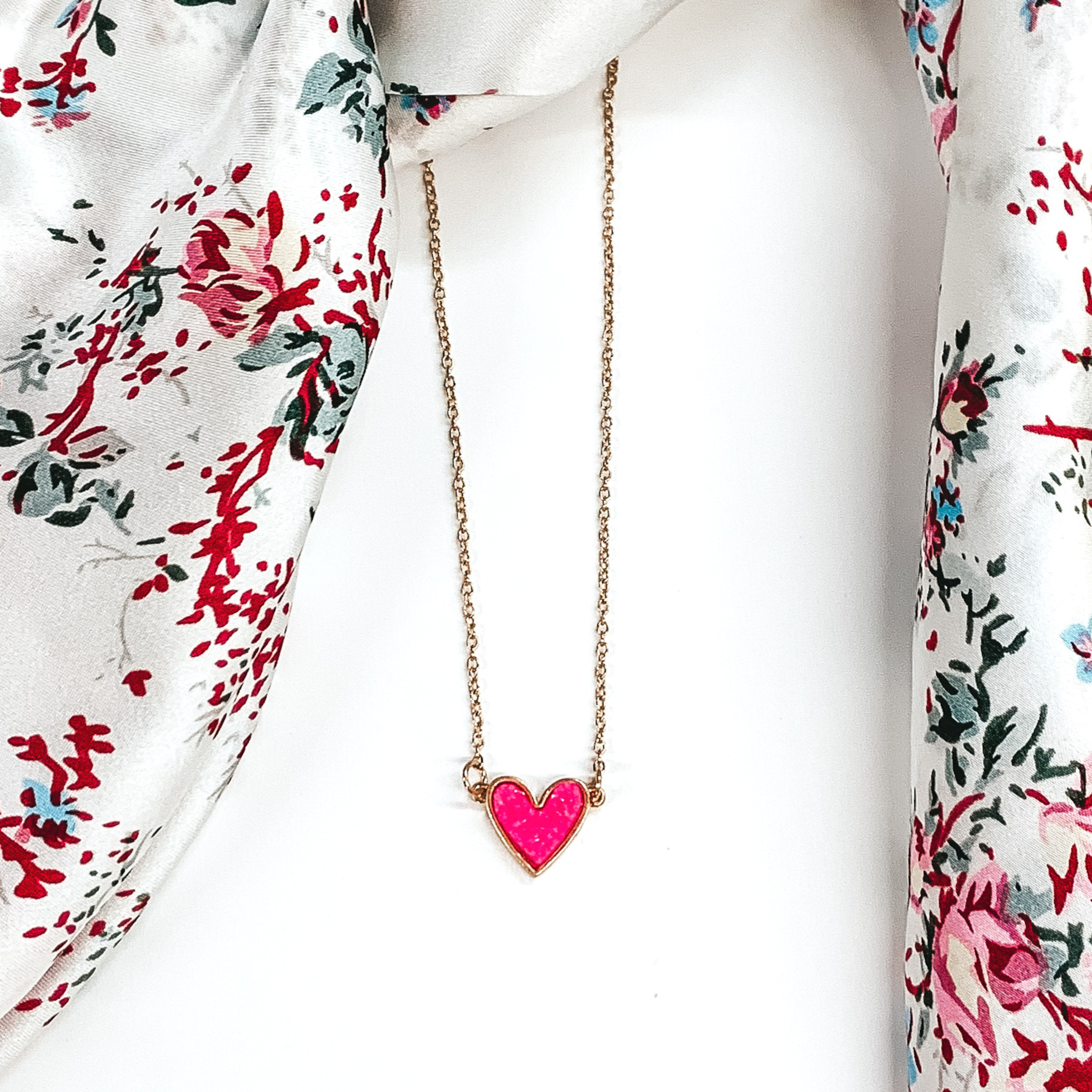 Small, gold chained necklace with a pink colored druzy heart pendant. This necklace is pictured on a white background with a white rag with colored flowers laying above the necklace.
