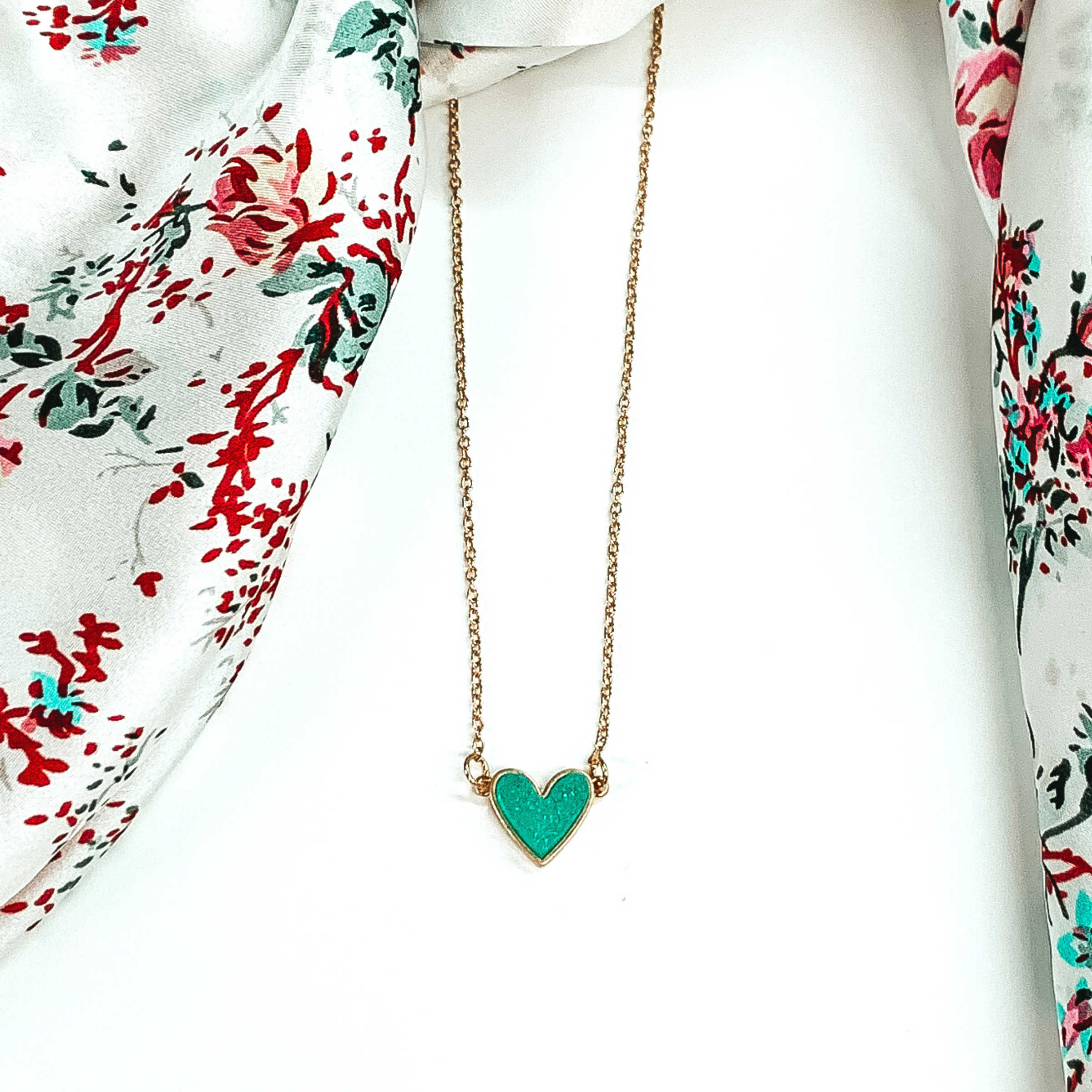 Small, gold chained necklace with a mint colored druzy heart pendant. This necklace is pictured on a white background with a white rag with colored flowers laying above the necklace. 