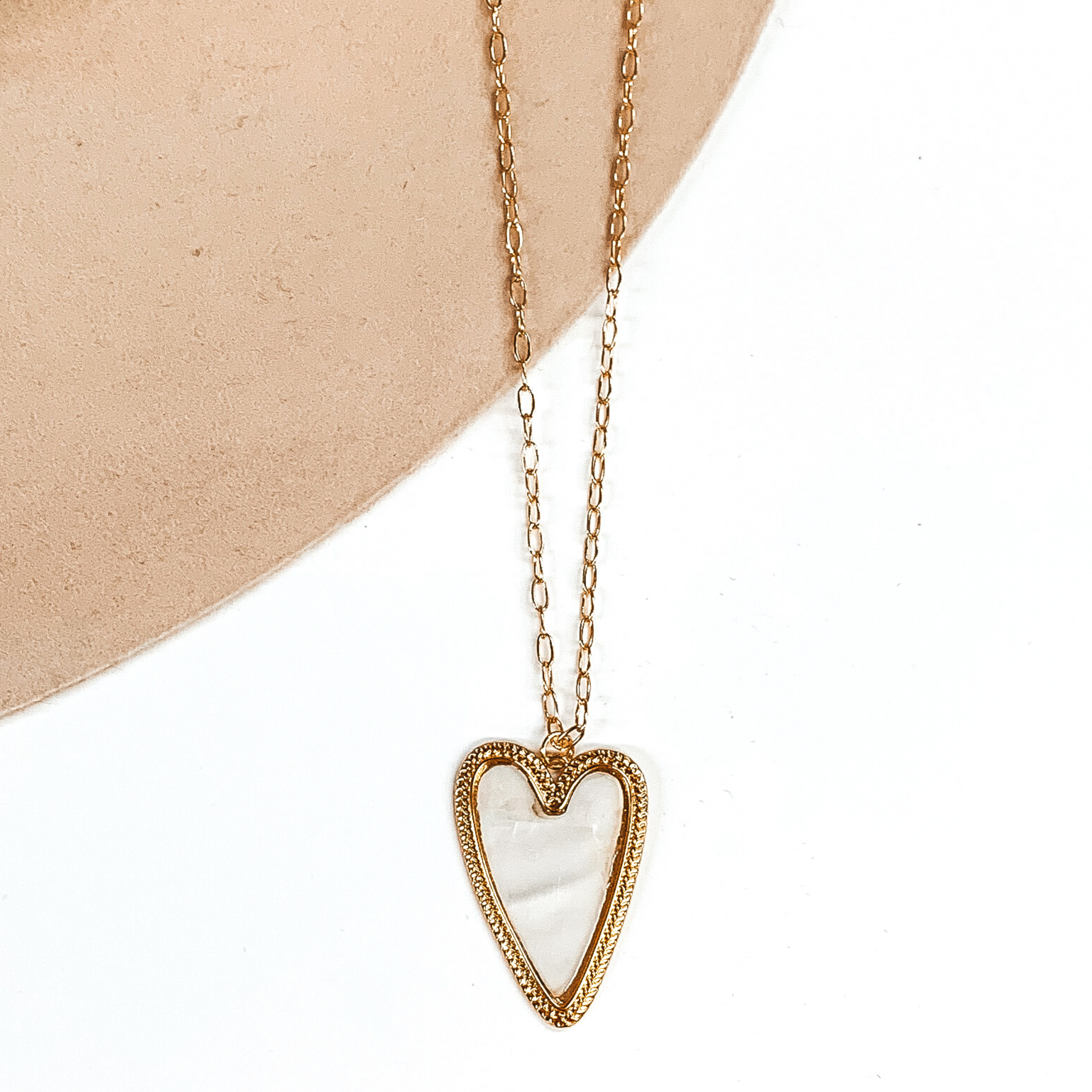 Thin gold chained necklace with heart pendant. The pendant is a gold, heart shaped pendant with a an ivory colored shell inlay. This necklace is pictured on a white and beige background. 