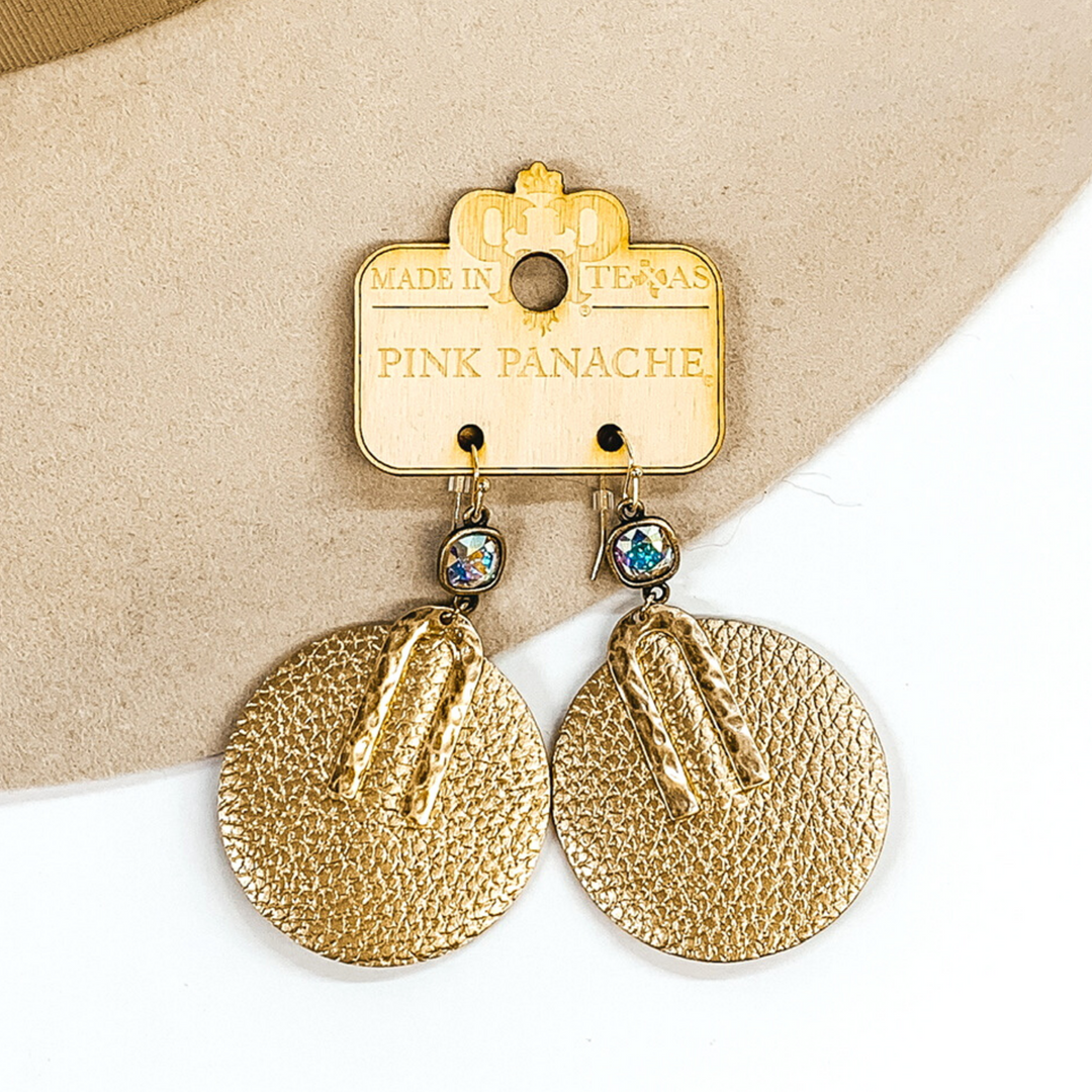 AB cushion cut crystal dangle earrings with a hanging, gold circle pendant. The pendant has a small gold "U" shaped accent. These earrings are pictured on a white and beige earrings.