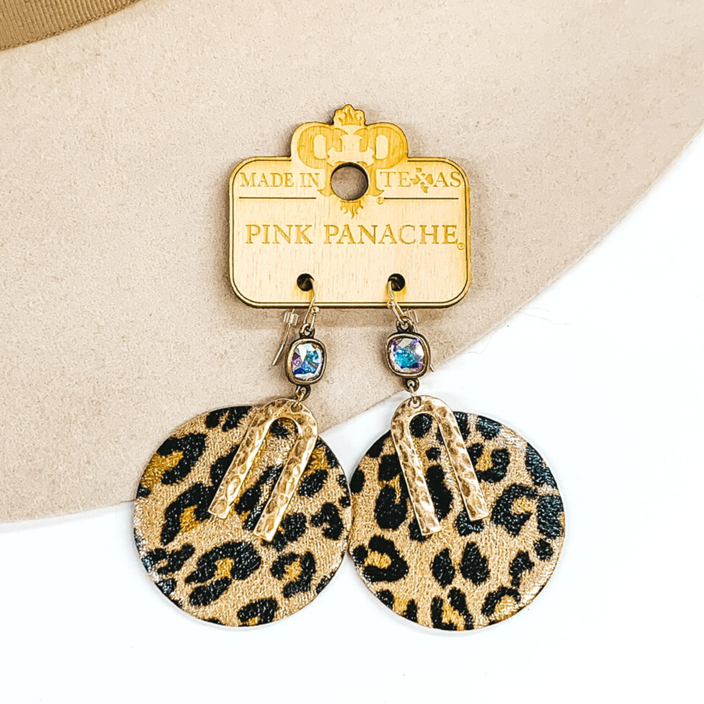 AB cushion cut crystal dangle earrings with a hanging, leopard circle pendant. The pendant has a small gold "U" shaped accent. These earrings are pictured on a white and beige earrings.