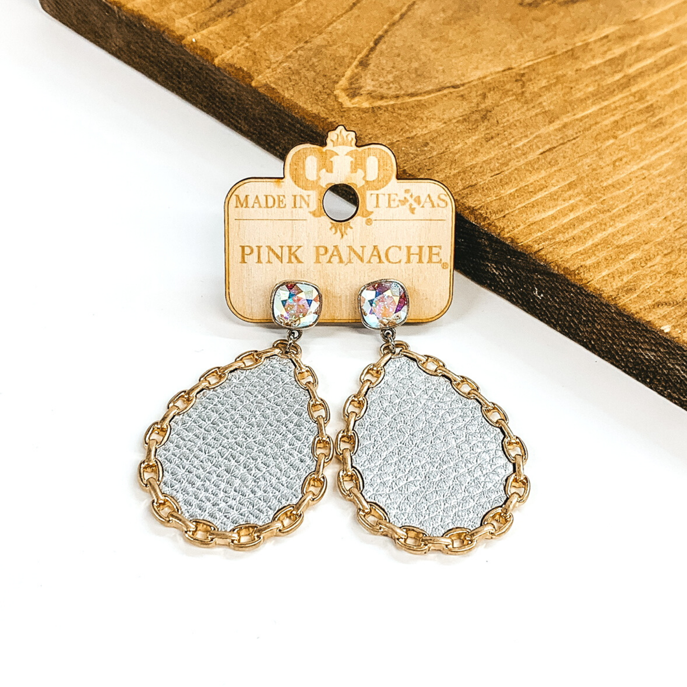 AB cushion cut crystal studs with a hanging teardrop pendant. The teardrop pendant is silver with a gold chain outline. These earrings are pictured on a white background with a piece of wood behind the earrings. 