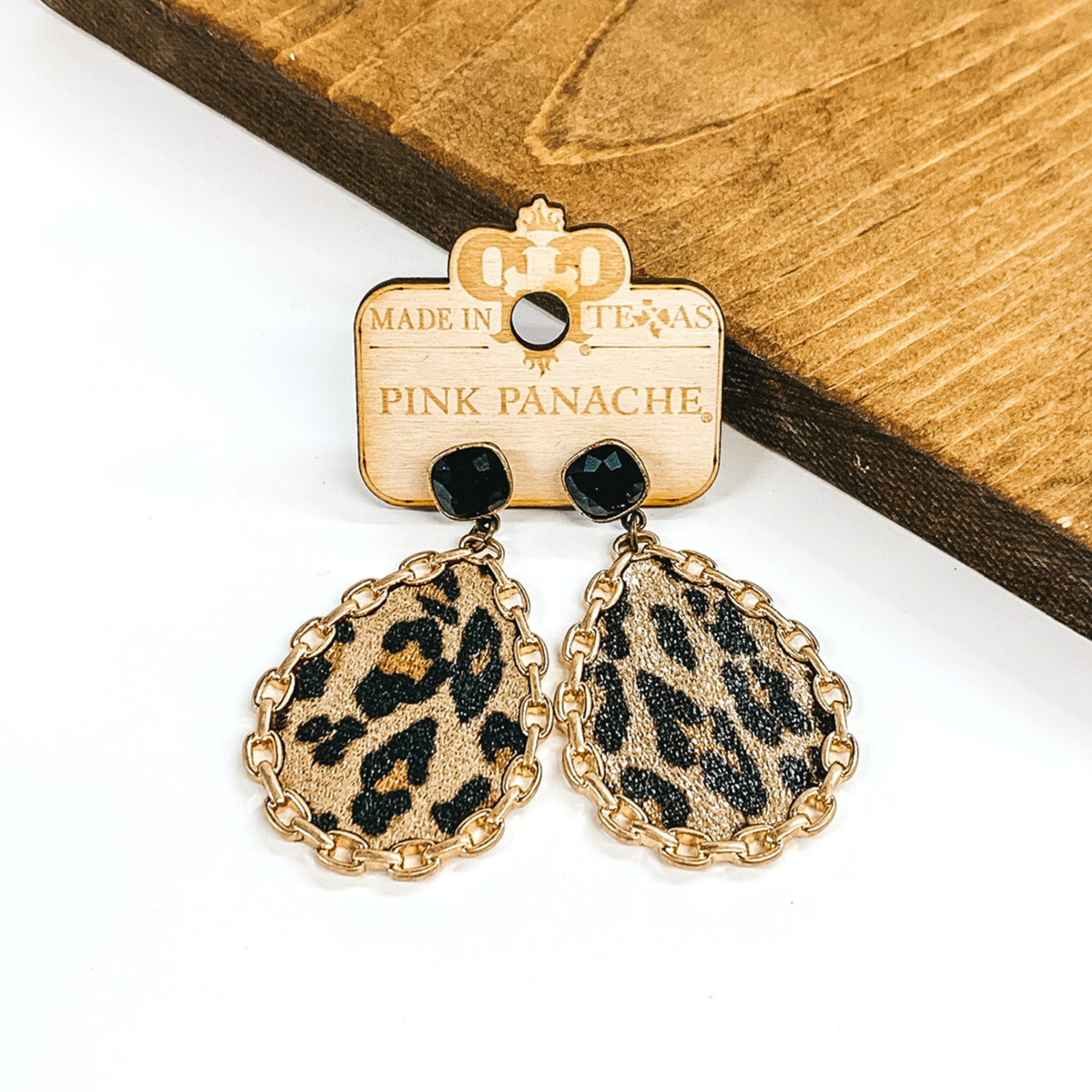 Black cushion cut crystal studs with a hanging teardrop pendant. The teardrop pendant is a leopard print with a gold chain outline. These earrings are pictured on a white background with a piece of wood behind the earrings.