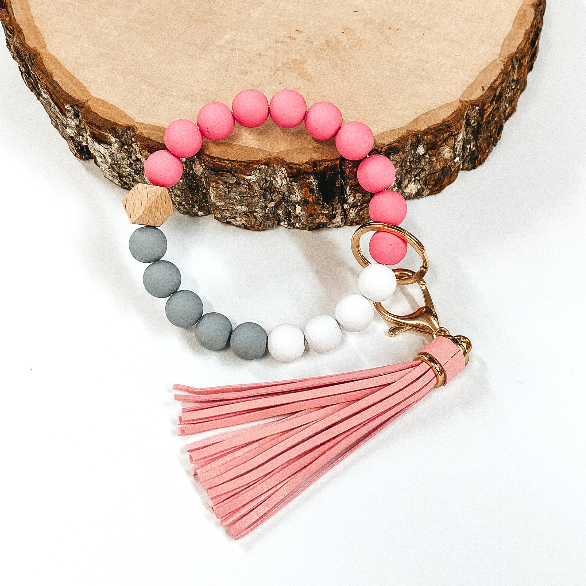 Matte beaded bangle key ring. The key ring includes pink, grey, and white. It also has a gold key ring and clasp with a pink tassel connected. This key ring is pictured laying on a piece of wood on a white background. 