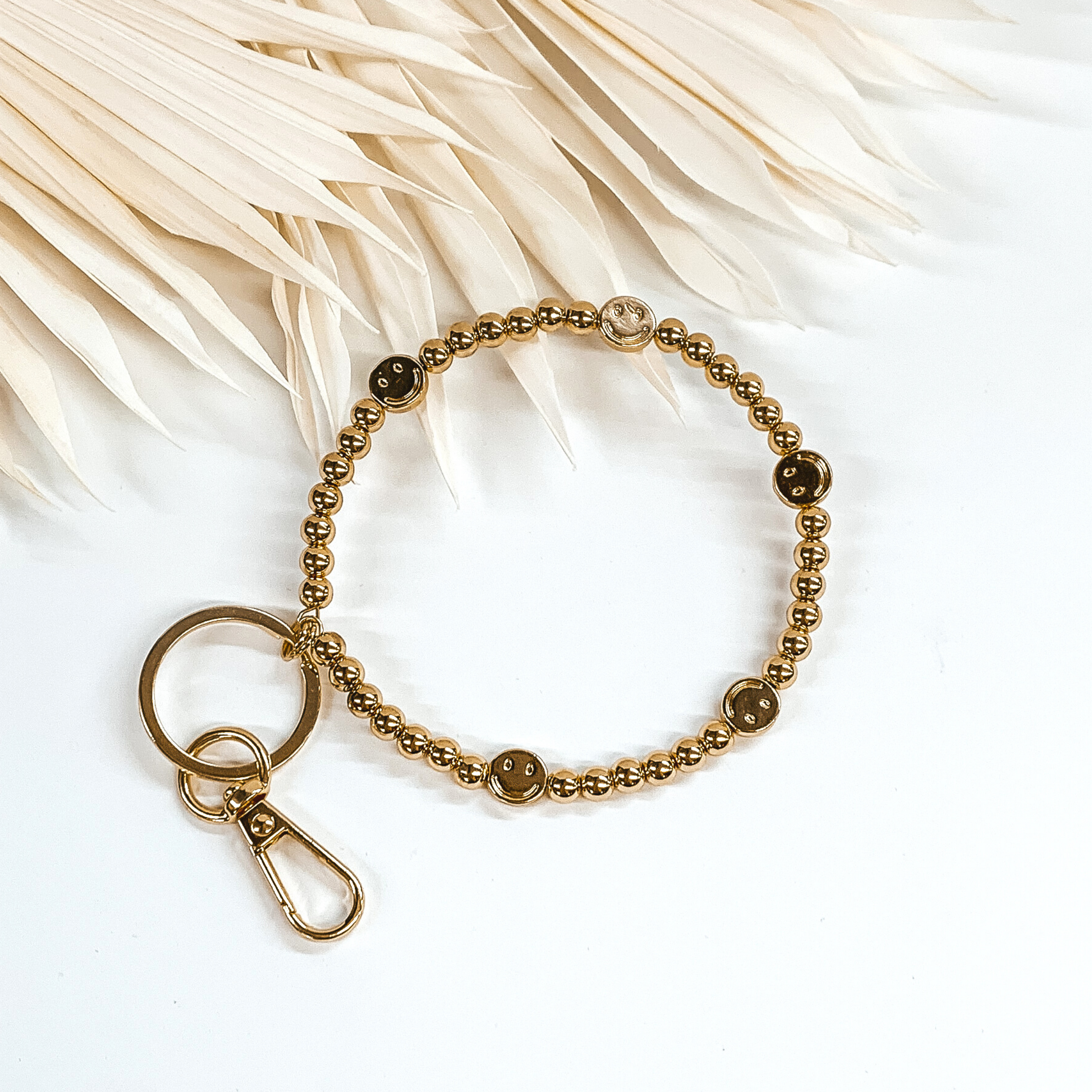 Gold beaded key ring with gold smiley face spacers. This key ring bracelet has a key ring and a clasp. This is pictured on a white background with white leaves at the top of the picture.
