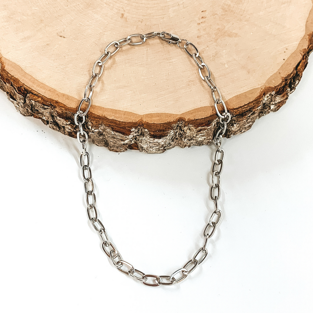 Silver paperclip chain in silver. This necklace is pictured laying partially on a piece of wood on a white background. 