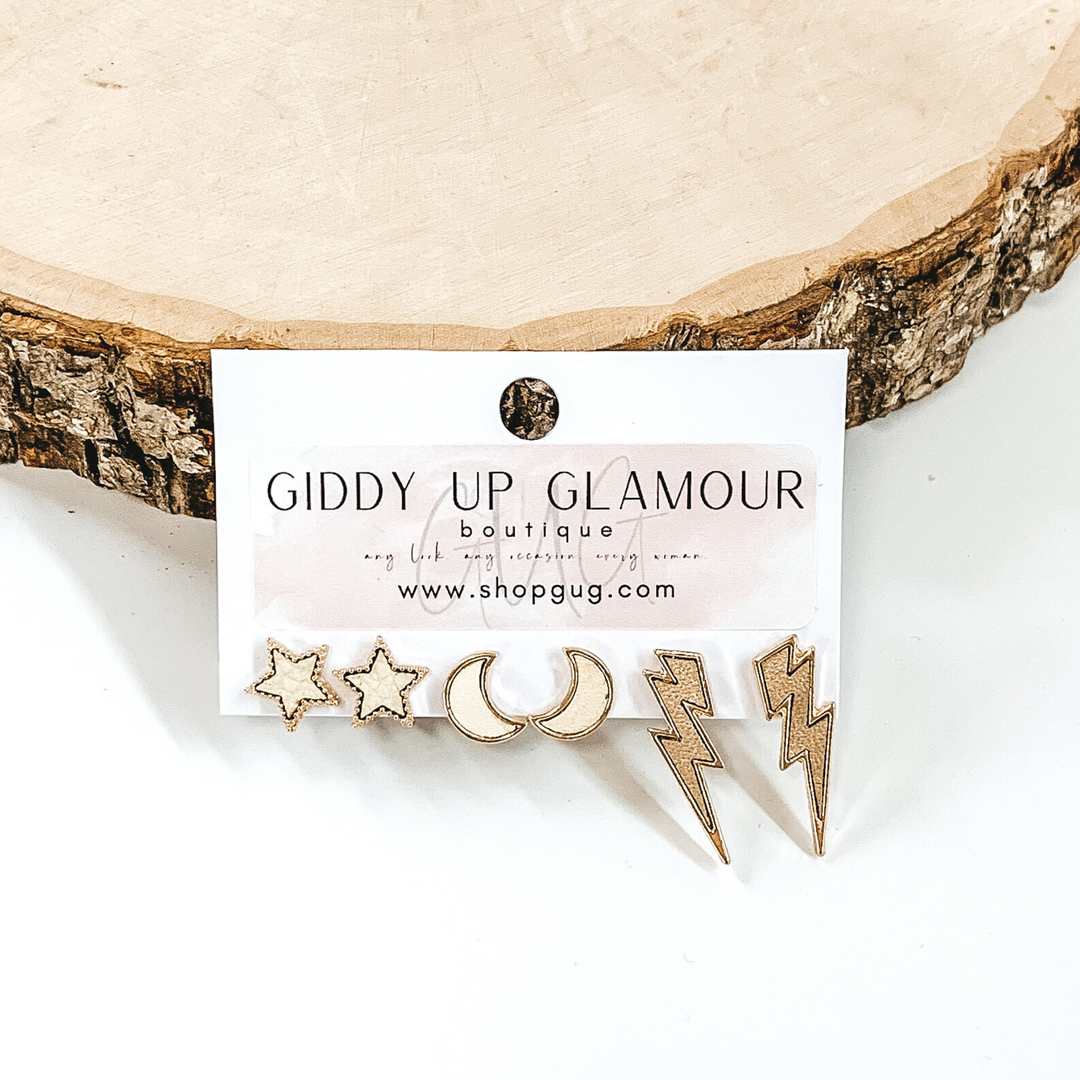 This set includes star studs, half moon studs, and lightning bolt studs. The stars are a dark ivory color, the moons are a bright ivory color, and the lightning bolt are a light tan color and all are outline in gold. This earring set is pictured on a white earrings holder in front of a piece of wood on a white background. 
