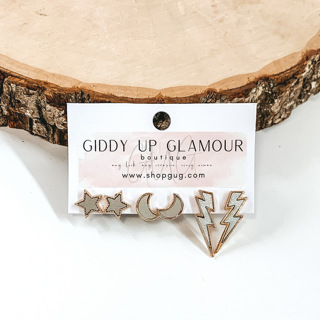 This set includes star studs, half moon studs, and lightning bolt studs. The studs are shades of light grey and all are outline in gold. This earring set is pictured on a white earrings holder in front of a piece of wood on a white background.