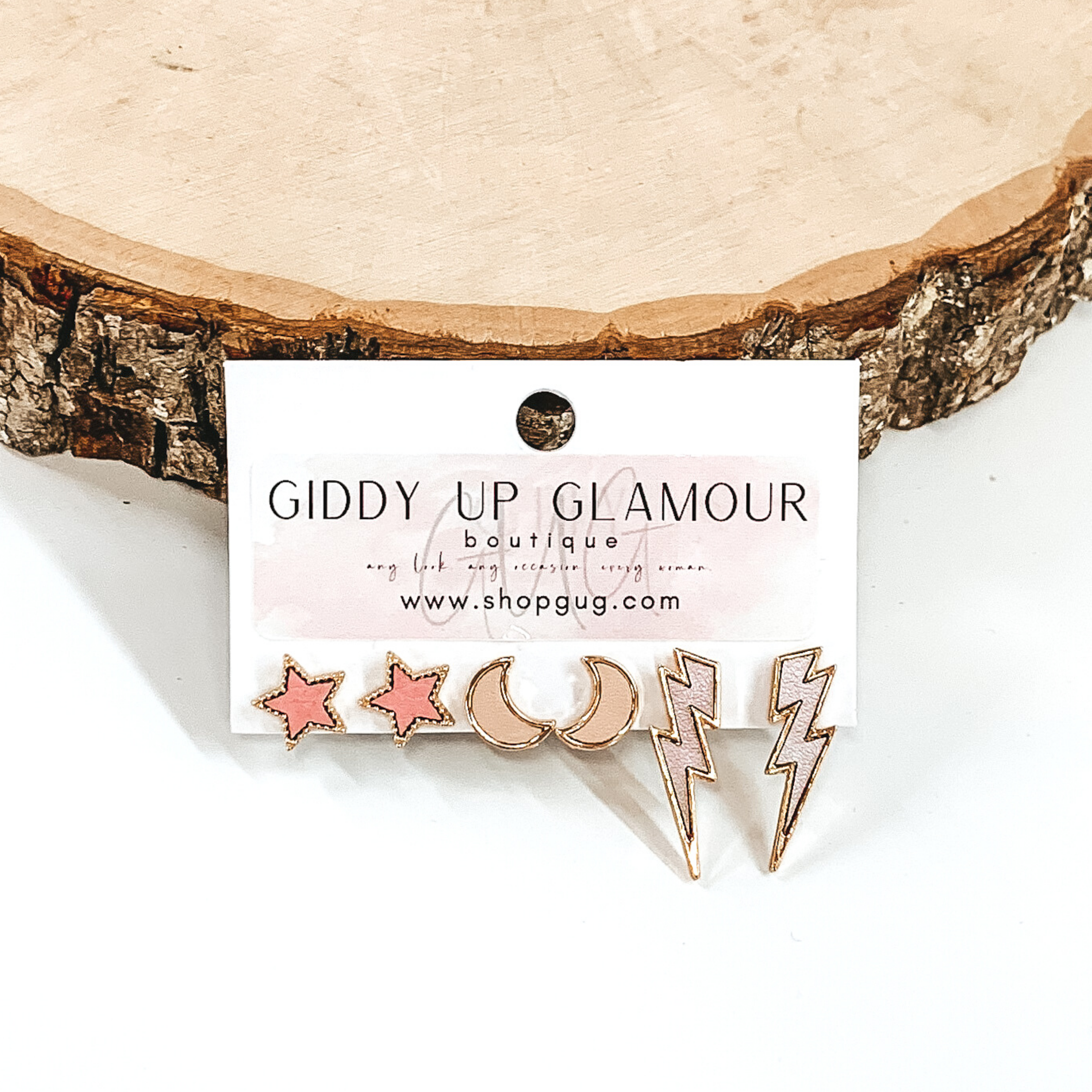 This set includes star studs, half moon studs, and lightning bolt studs. The stars are a light pink color, the moons are a dark pink/ivory color, and the lightning bolts are a light pink/lilac color and all are outline in gold. This earring set is pictured on a white earrings holder in front of a piece of wood on a white background.